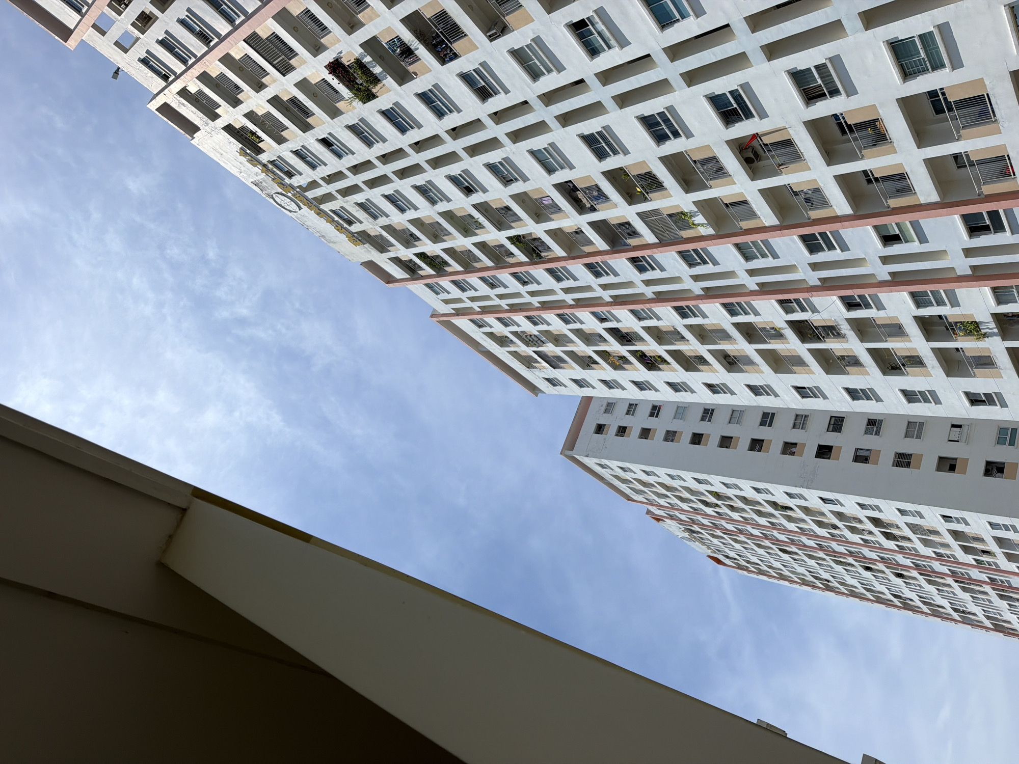 The image shows two tall apartment buildings against a bright blue sky with scattered clouds. The buildings are predominantly white with pink accents along the vertical edges of balconies. They feature a grid-like pattern of windows and balconies. The building on the right is closer and more detailed, showing individual units with balconies, windows, and some with air conditioning units or plants. The building on the left is partially obscured by another structure in the foreground on the left side of the image. A golden text inscription is visible on the facade of the right building, though it is partially cut off. The angle of the shot is looking upwards towards the sky, emphasizing the height of the buildings. The scene depicts an urban residential environment, likely in Bac Nha Trang Ward, Vietnam, given the architectural style and tropical sky. No people are clearly visible in the image.