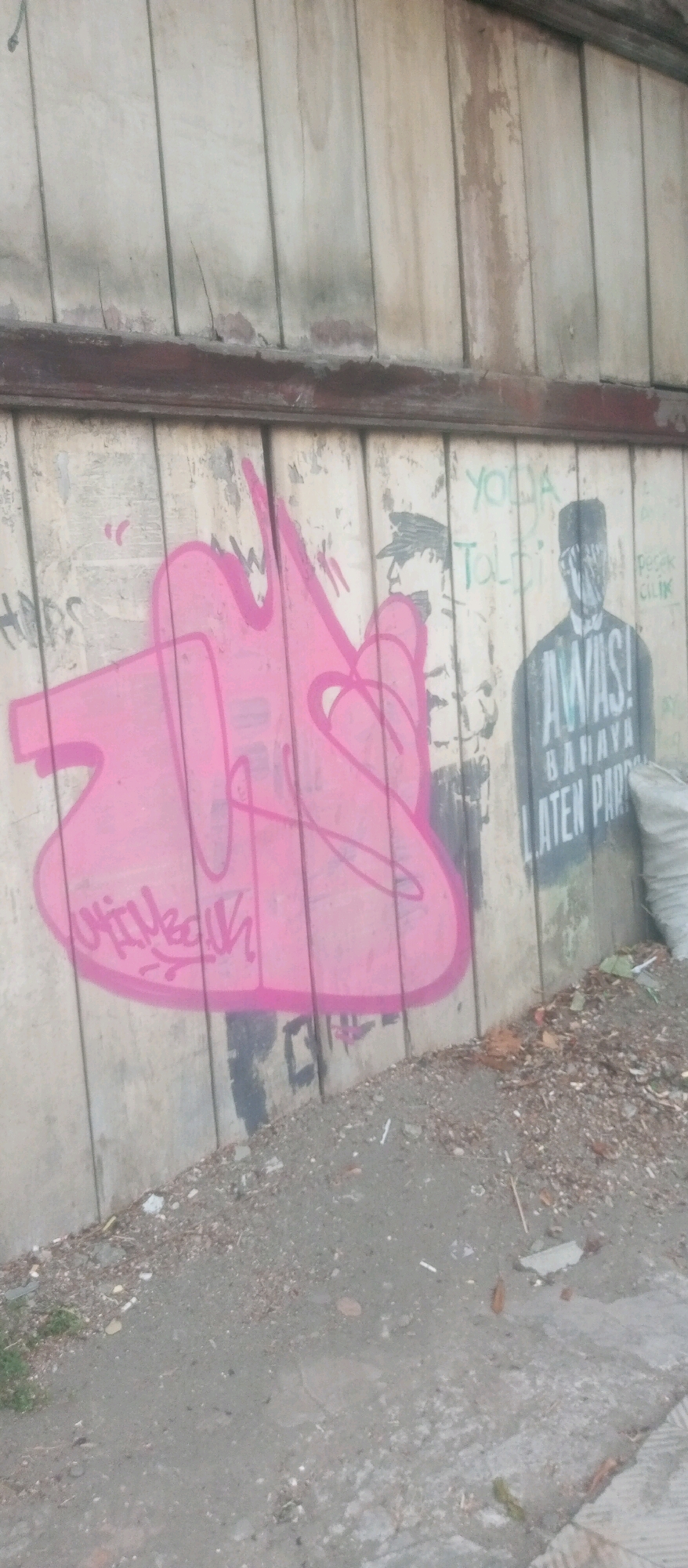 The image captures an exterior wall in Kediri City, Indonesia, adorned with graffiti. On the left, a large, stylized pink tag dominates the surface. To the right of the pink graffiti, two black and white stenciled figures are visible. The first figure on the left appears to be a person in a hat. The second figure on the right is dressed in a suit and a fez-like hat, with text superimposed on it. The text includes "AWAS! BAHAYA LATEN PART" written vertically. In the upper right portion of the wall, some green graffiti reads "YODA" and "TOLERAN". The wall itself is made of vertical wooden planks. The ground in front of the wall is covered with dirt, debris, and some discarded materials, including a white bag partially visible on the far right. The overall scene depicts an urban environment with visible signs of vandalism and decay.