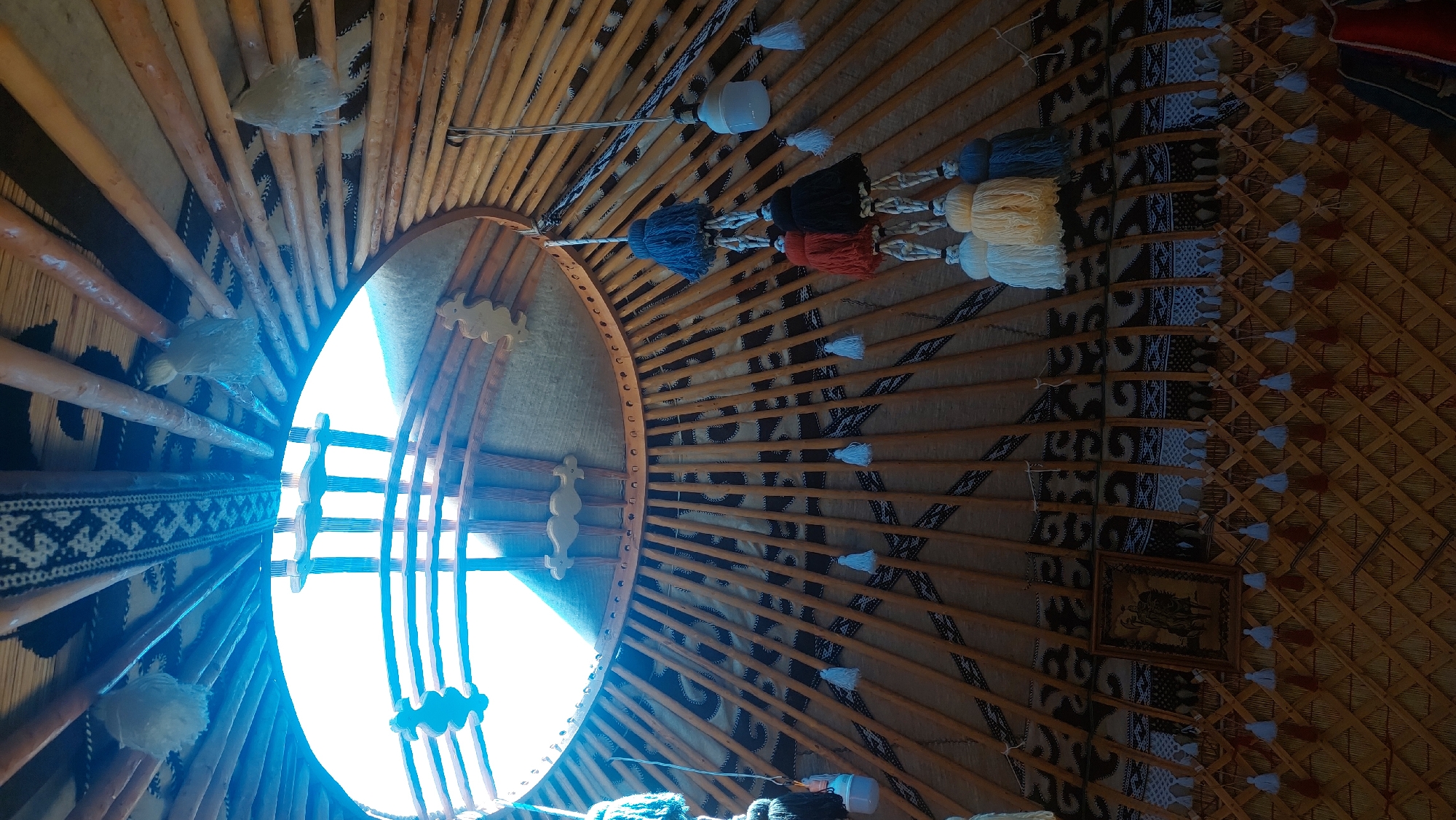 This is a low-angle, interior shot of a traditional yurt, likely located in Бактуу-Долоноту, Kyrgyzstan. The domed ceiling, characteristic of yurts, is visible, with numerous wooden poles radiating from the central opening, which is letting in bright daylight. Decorative textiles with geometric patterns are draped along the upper interior walls and ceiling. Several fabric tassels and a small framed artwork are hanging from the poles. The walls are constructed from a lattice of wooden poles covered by a woven material. The overall impression is of a rustic, culturally rich interior space. There are no people visible in the image.