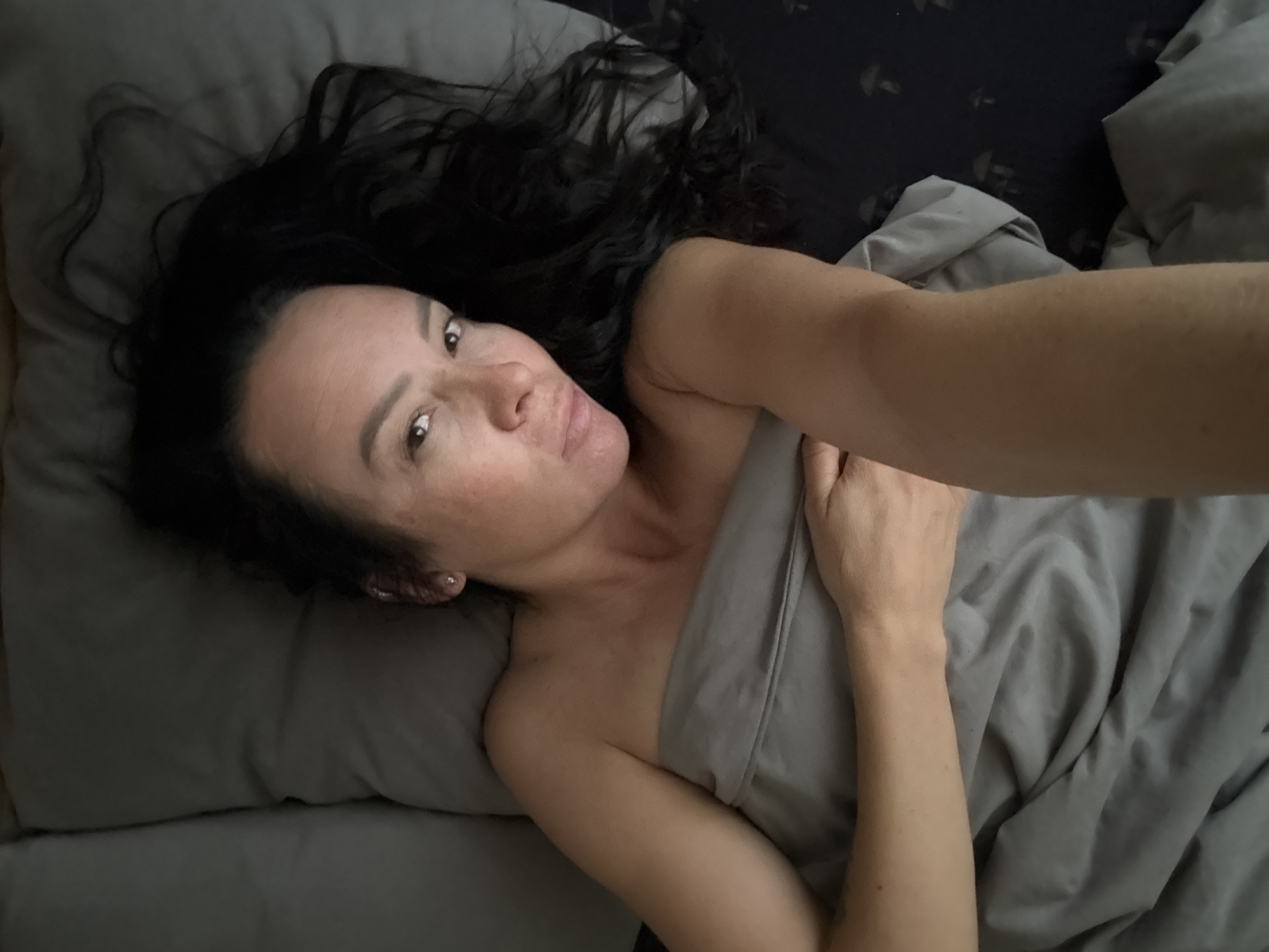 A woman with long, dark hair is lying in bed. She is under a gray blanket, with her left arm resting on top of it, holding it in place. Her right arm is extended towards the camera. She is looking directly at the camera with a neutral expression. The bed is made with gray pillows and a dark blue sheet with a mushroom pattern. The scene appears to be indoors, likely a bedroom.
