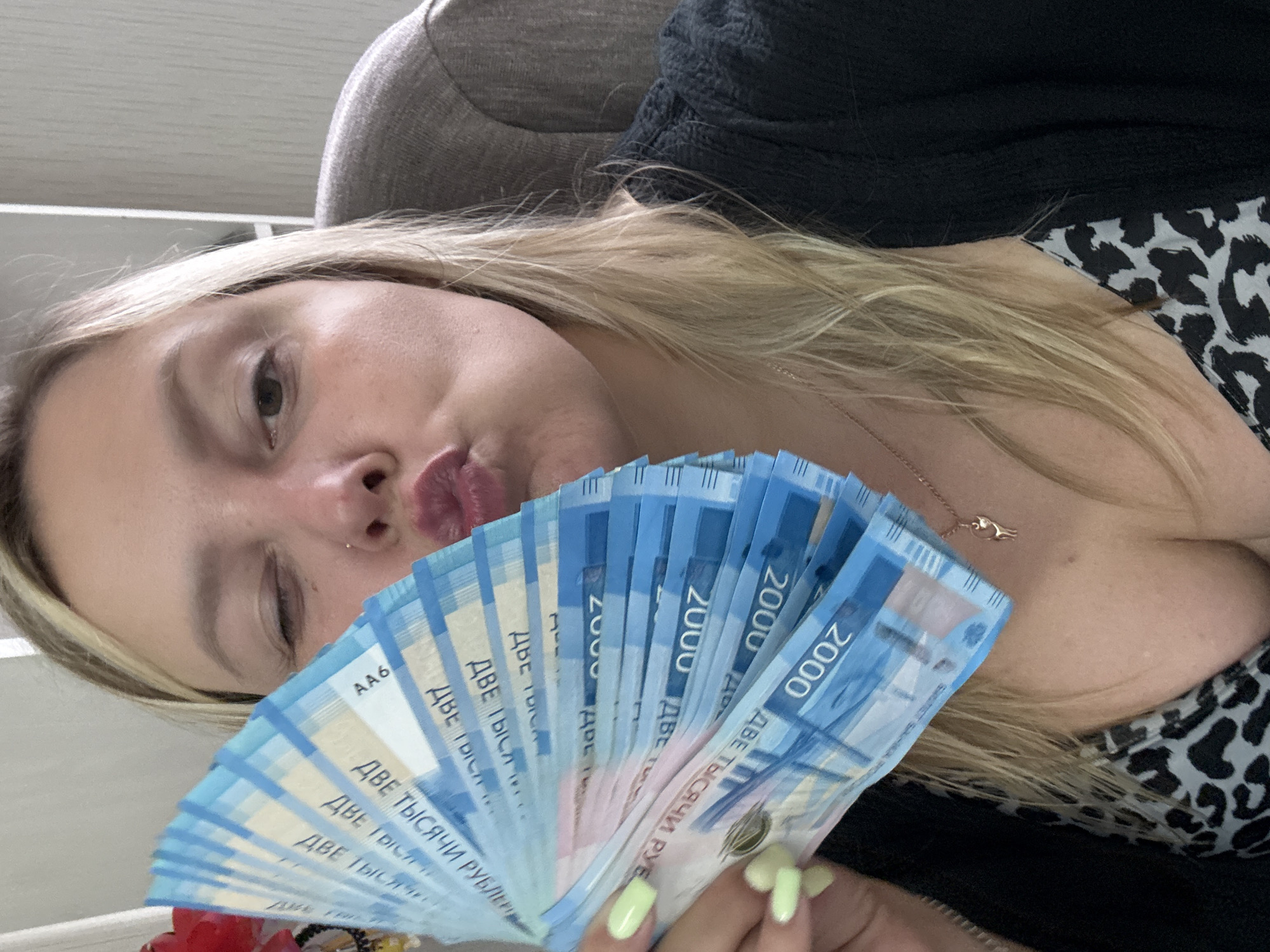 A blonde woman with light skin and plump lips is holding a fan of Russian 2000 ruble banknotes. She is winking, and her head is tilted to the left. The woman is wearing a black sweater over a leopard-print top and a thin necklace with a small pendant. Her fingernails are painted a light green color. The background is blurred and appears to be an indoor setting with a gray chair visible. The image does not provide any specific information about the location in the city of Noviy, Russia.