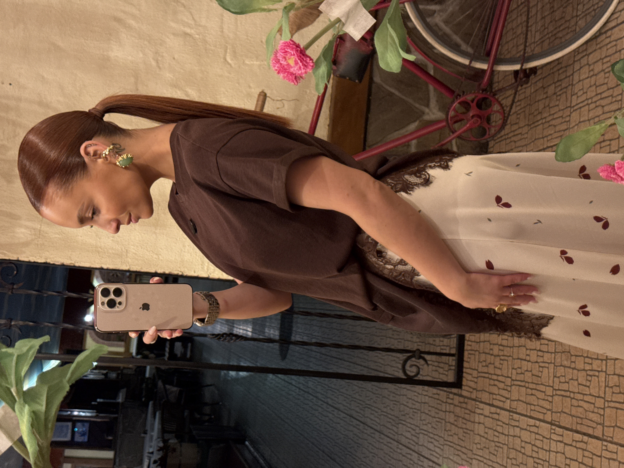 A woman with reddish-brown hair pulled back into a ponytail is holding a gold iPhone 14 Pro and is positioned indoors, possibly in a cafe or restaurant. She is wearing a brown, loose-fitting top and a patterned dress with lace trim. The environment includes interior details such as tile flooring, ironwork or railings, and plants. A red bicycle with pink flowers is visible in the background. There's no verifiable evidence to confirm the location is Chaikovsky, Russia, based solely on the image.