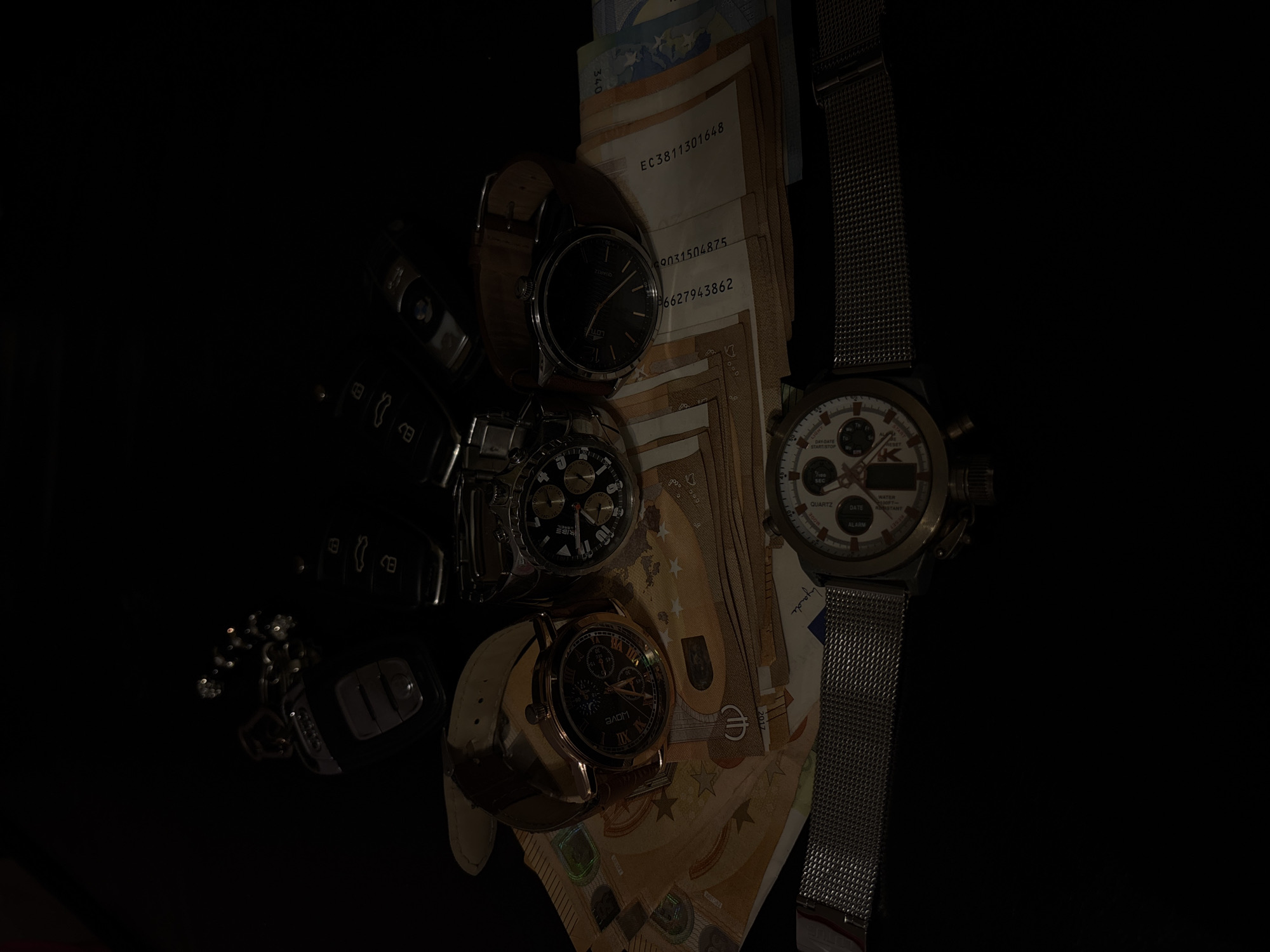The image displays a dark, top-down still-life arrangement of luxury and personal items on a black surface, likely indoors in Dolni Tsibar, Bulgaria. The scene is lit dramatically, with most details emerging from deep shadow.
The primary subjects are four wristwatches, several Euro banknotes, and multiple car keys or fobs.
The watches vary in style:
1. One features a brown leather strap and a black dial with "LOTU" branding.
2. Another is a silver-toned watch with a black dial and chronograph subdials.
3. A rose gold-toned watch has a brown leather strap, a black dial with Roman numerals, and chronograph functions, bearing "WOVE" branding.
4. The most prominent watch is silver-toned with a mesh strap, a light-colored dial, chronograph subdials, and a digital display. Visible text on this watch includes "QUARTZ", "DATE", "WATER RESISTANT", "ALARM", "START/STOP", "RESET", and "LIGNT" (possibly part of "LIGHT").
Several Euro banknotes, appearing to be 50 Euro notes, are fanned out beneath the watches. Visible text and serial numbers on these notes include "EC3811301648", "69031504875", "6627943862", "340", and the year "2017". A signature or stylized text "you jande" is also visible on one note.
Multiple car keys and fobs are positioned to the left, largely obscured by shadow, with one key resembling a BMW design. The composition evokes themes of wealth, personal belongings, and time.