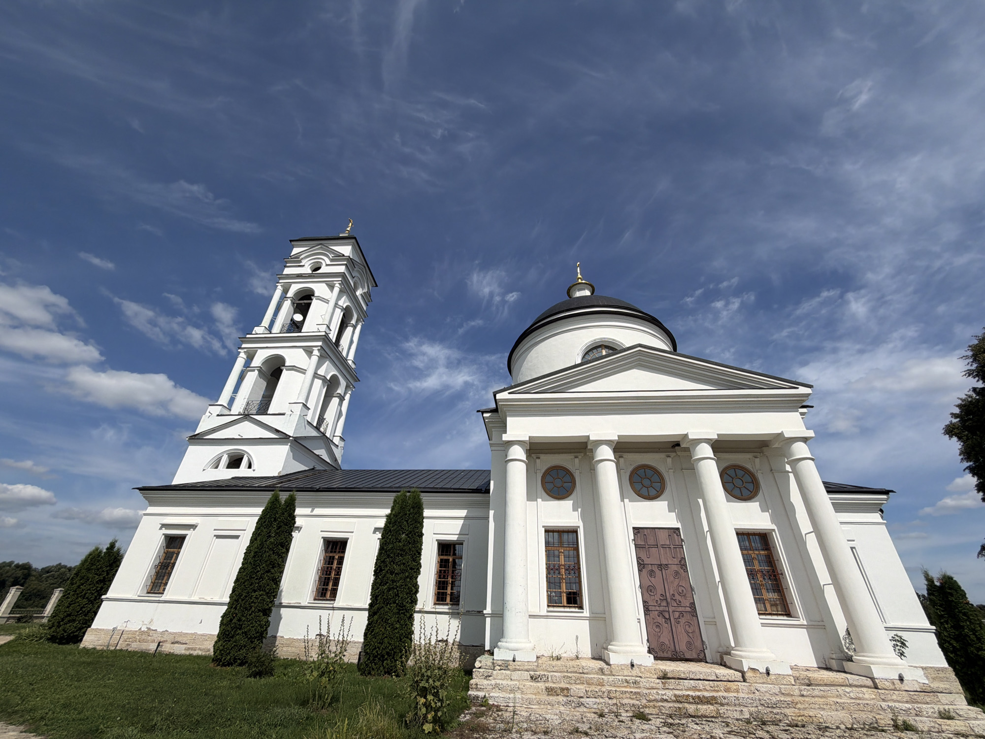 A wide-angle shot captures the Church of the Assumption of the Most Holy Mother of God in Skoryakovo, Russia. The white stone church features a prominent bell tower with arched openings and a golden cross topping its roof. Adjacent to the bell tower, a domed main building with classical columns and a large wooden door is visible. The structure is surrounded by green grass and tall, slender evergreen trees, set against a backdrop of a bright blue sky with wispy white clouds. The scene is a bright, clear day.