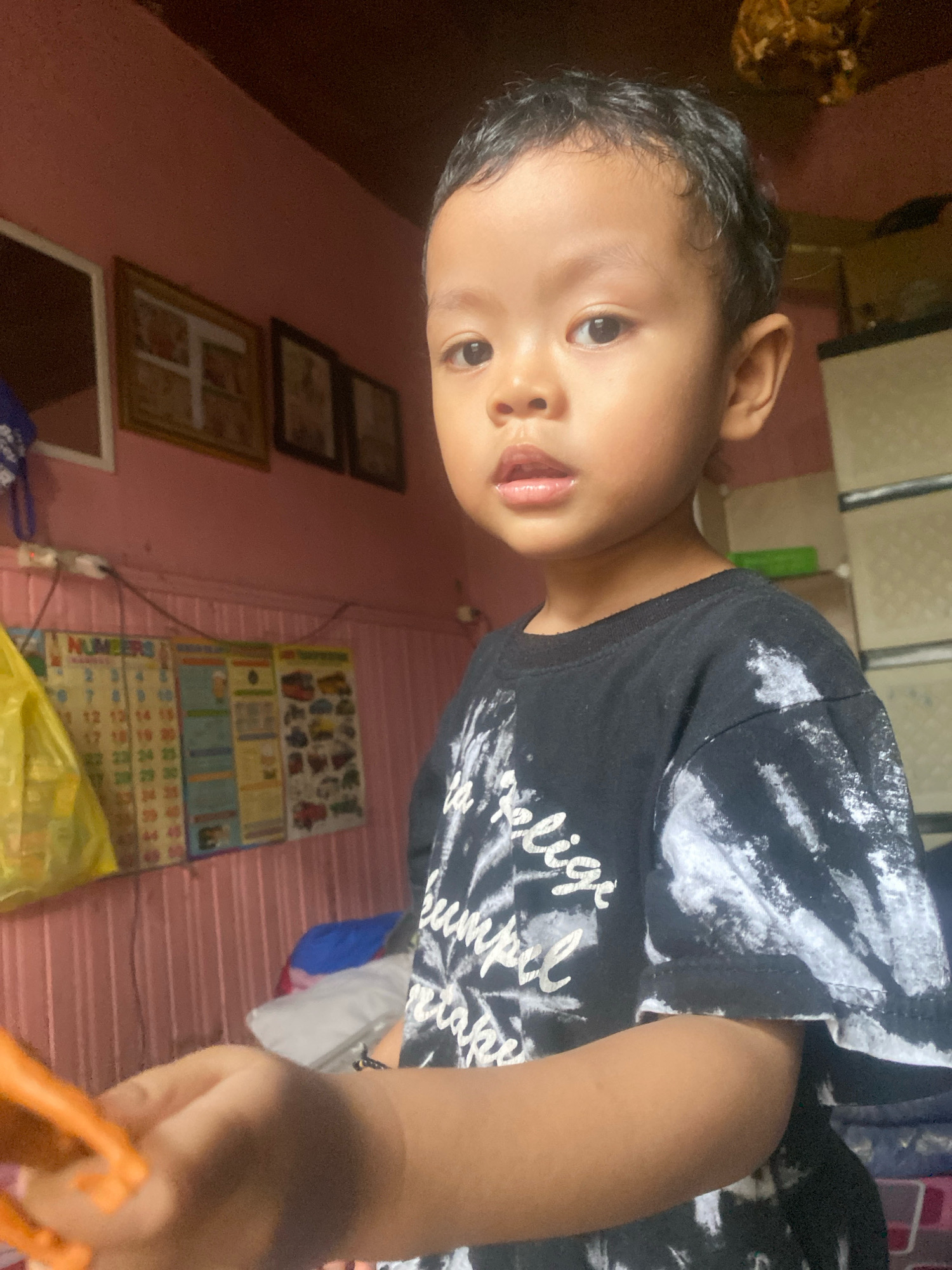 A young boy is shown from the chest up, looking towards the camera with a neutral expression. He has dark hair and is wearing a black t-shirt with a white, tie-dye-like pattern and white text. The background appears to be a domestic interior, possibly a bedroom, with a pink wall. On the wall behind the boy, there are several framed pictures and a colorful chart with the word "NUMBERS" visible. A yellow plastic bag hangs on the left side of the frame. The lighting suggests an indoor setting during the day. The image was taken in Banjarmasin, Indonesia.