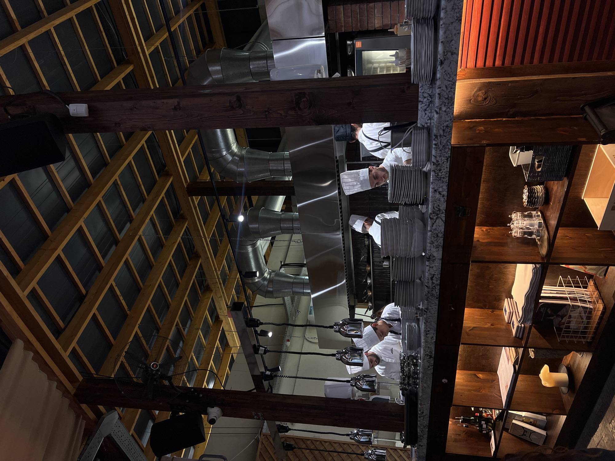 The image shows a kitchen in a restaurant located in Yerevan, Armenia. Four chefs in white uniforms and hats are working in a professional kitchen under a large metal vent hood. A stack of white plates is on the countertop. Exposed wooden beams support the ceiling, along with ventilation ducts and overhead lighting fixtures. Shelves are visible on the right side, holding various items.