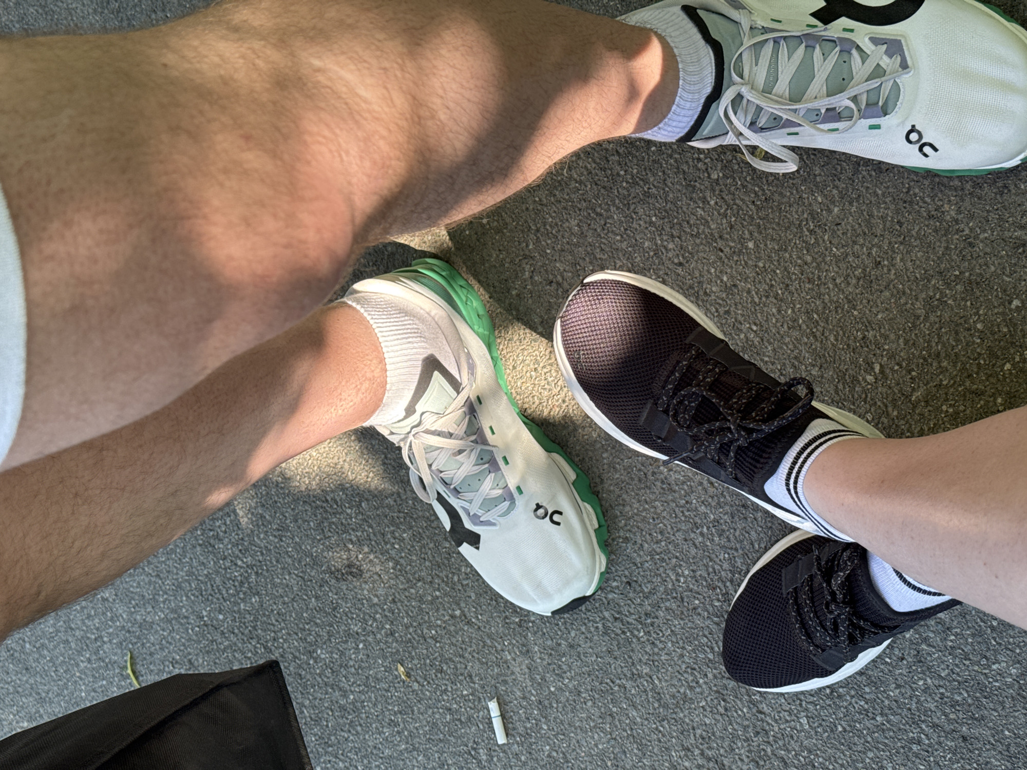 The image presents a high-angle view of the lower legs and feet of two individuals positioned on a grey asphalt surface, indicative of an outdoor urban setting. Strong direct sunlight illuminates the scene, casting distinct shadows across the pavement.
In the foreground and to the left, one individual's legs are crossed, wearing black mesh athletic shoes with white midsoles and black laces. White ankle socks with two thin black horizontal stripes at the cuff are visible. The skin on these legs is light-toned and relatively hairless.
To the right and background, the lower legs of a second individual are depicted. This individual wears white 'On Running' brand athletic shoes, featuring a black 'oQ' logo on the side, prominent green elements on the midsole and outsole, and subtle light green/grey and purple accents around the white laces. White ribbed ankle socks are worn. The skin on these legs is medium-toned and displays a noticeable amount of body hair.
A discarded cigarette butt with a filter and a faint colored band is visible on the asphalt near the toe of the white 'On' shoe. In the upper left quadrant, a portion of a dark fabric bag rests on the ground, alongside a small, light green leaf. The asphalt surface and urban context are consistent with a location such as Riga, Latvia.