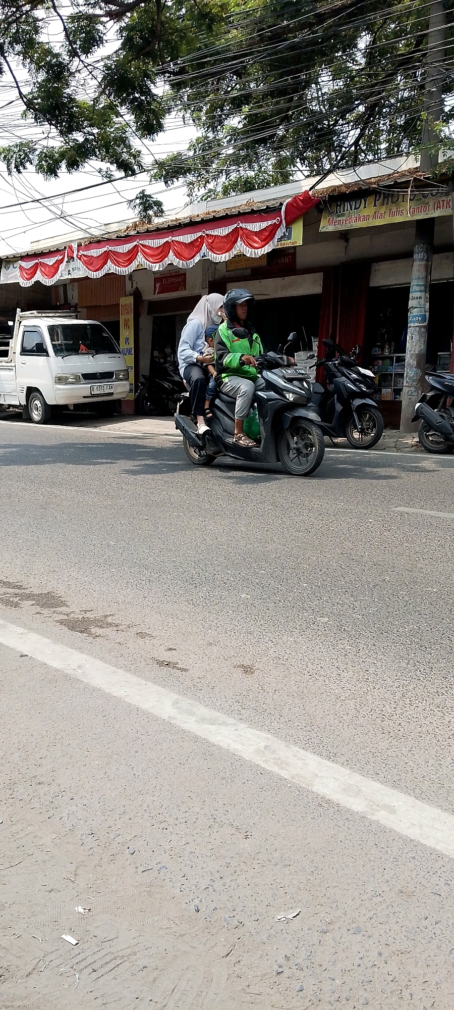 In Karangsatria, Indonesia, a family of three rides a dark-colored scooter. A driver in a green jacket and black helmet operates the vehicle. Next to the driver, a woman wearing a light blue long-sleeved shirt and a white hijab holds a young child. The child is dressed in a light-colored shirt. They are on a paved road with a white line marking the lane. In the background, a white pickup truck is parked near a building with a red and white awning. Several other parked motorcycles are visible. The scene is illuminated by daylight, with trees and overhead wires contributing to the urban landscape.