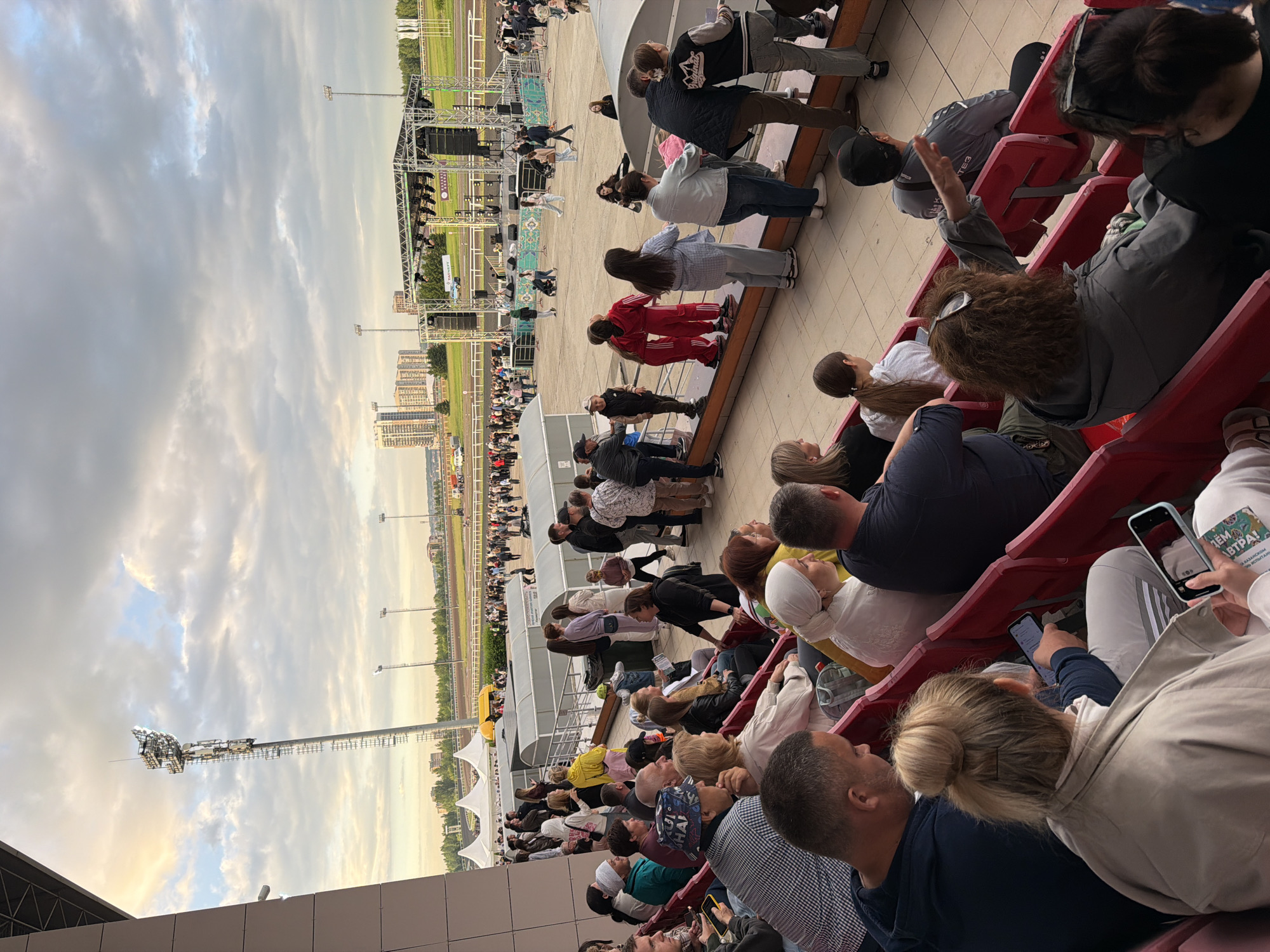 The image shows an outdoor event with a large crowd gathered in stadium seating, facing a stage and a field. The sky is cloudy with some hints of sunlight. A tall lighting tower is visible in the background. The people in the foreground are seated in red stadium seats, and many appear to be looking towards the stage. Some individuals are holding phones and taking pictures or videos. The overall atmosphere suggests a concert or a large public gathering.
