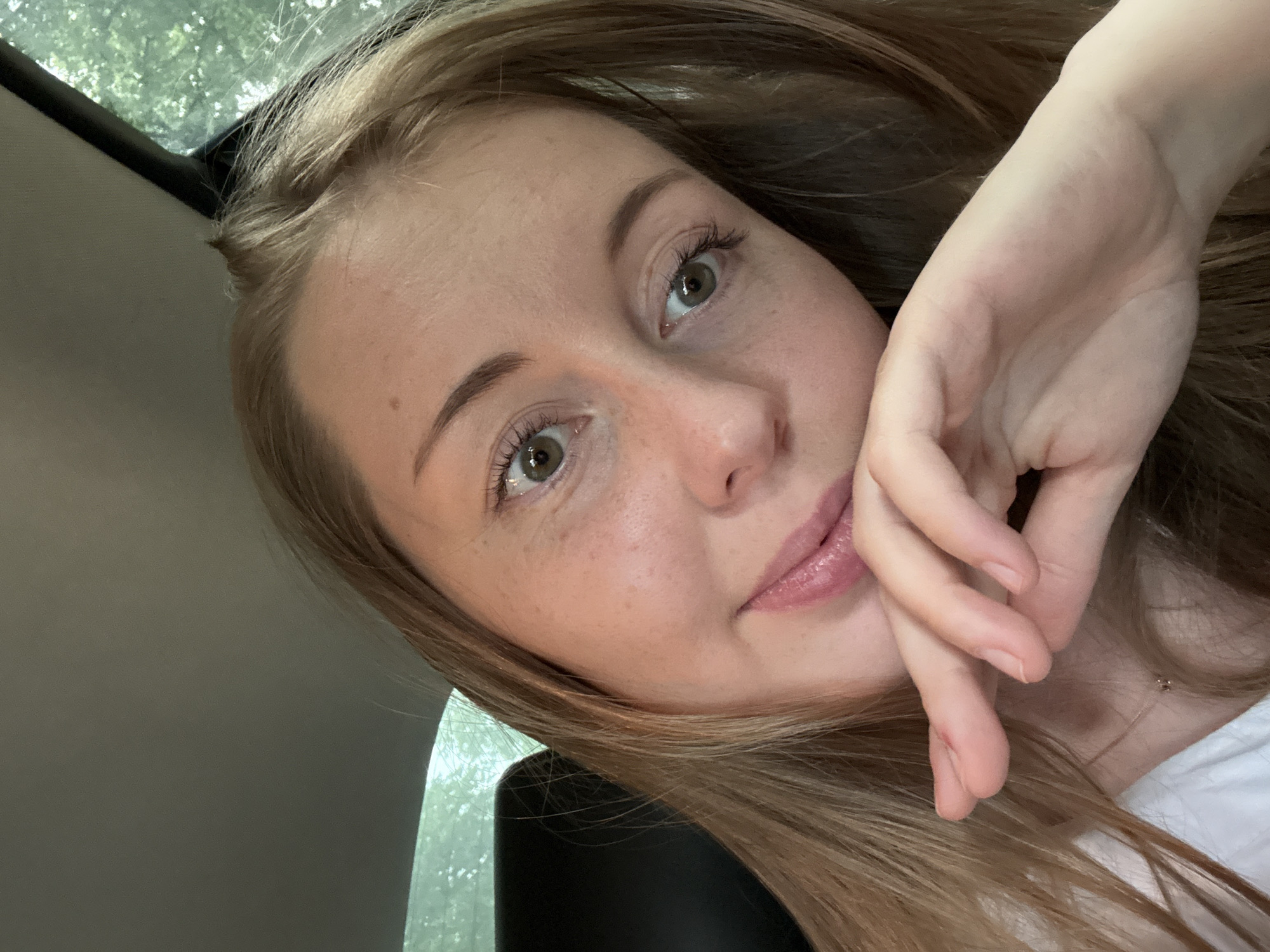 A young woman with blonde hair and green eyes is pictured from the chest up in a car. She is wearing a white shirt and a delicate necklace. Her right hand is touching her chin, and her head is tilted to the left, creating a playful and intimate pose. The background is blurred, showing glimpses of trees through the car window, suggesting an outdoor, possibly natural setting. The lighting is soft and natural, highlighting the woman's freckled skin and rosy cheeks.