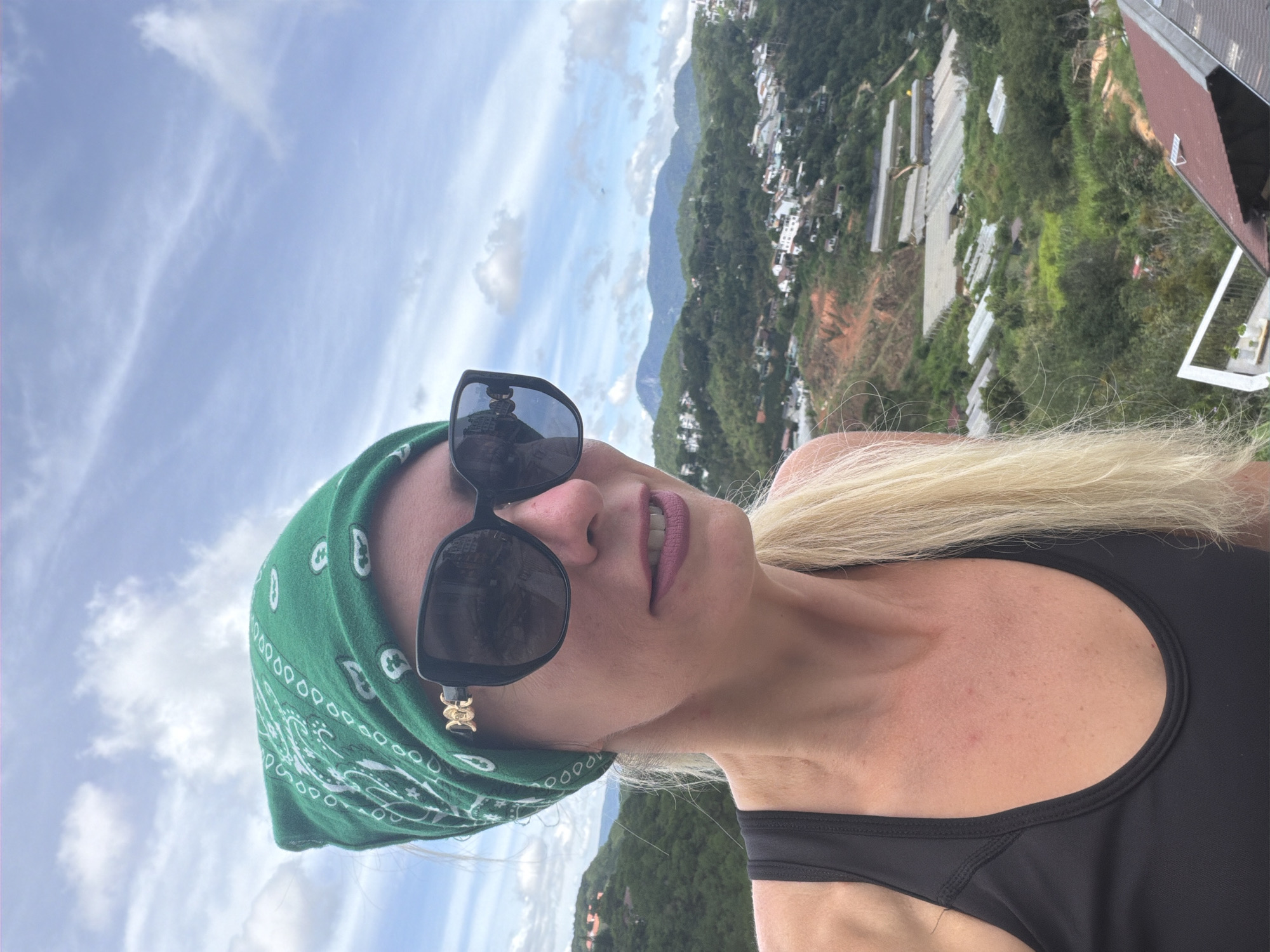 A blonde woman with fair skin wears a green bandana and large black sunglasses with gold accents. She is wearing a black tank top. The woman is facing right and looking forward with her mouth slightly open, revealing her teeth. Her blonde hair extends past her shoulders. Behind her, the landscape of Da Lat, Vietnam, unfolds. The scene features rolling green hills dotted with white buildings and greenhouses. The sky is partly cloudy with a mix of blue and white.
