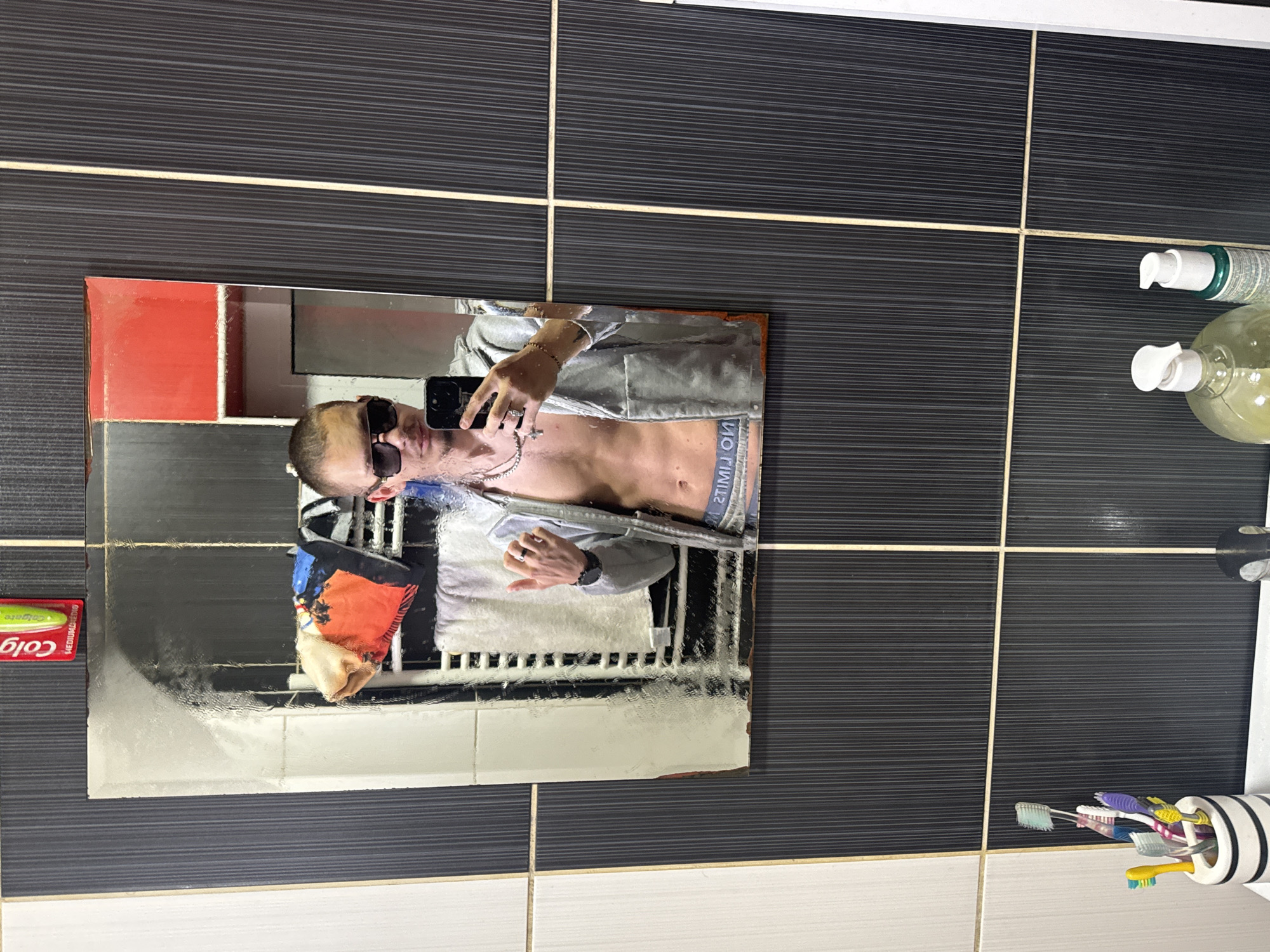 The image depicts a bathroom setting. A man with sunglasses is reflected in a mirror, appearing to take a photo of himself. He is wearing an unzipped silver jacket, revealing his torso and the waistband of his underwear. In the reflection, a patterned towel hangs behind him. The bathroom features a mix of black and white tiled walls, with visible toiletries such as soap dispensers and toothbrushes. There is no indication that this image was taken in Vetren Dol, Bulgaria.