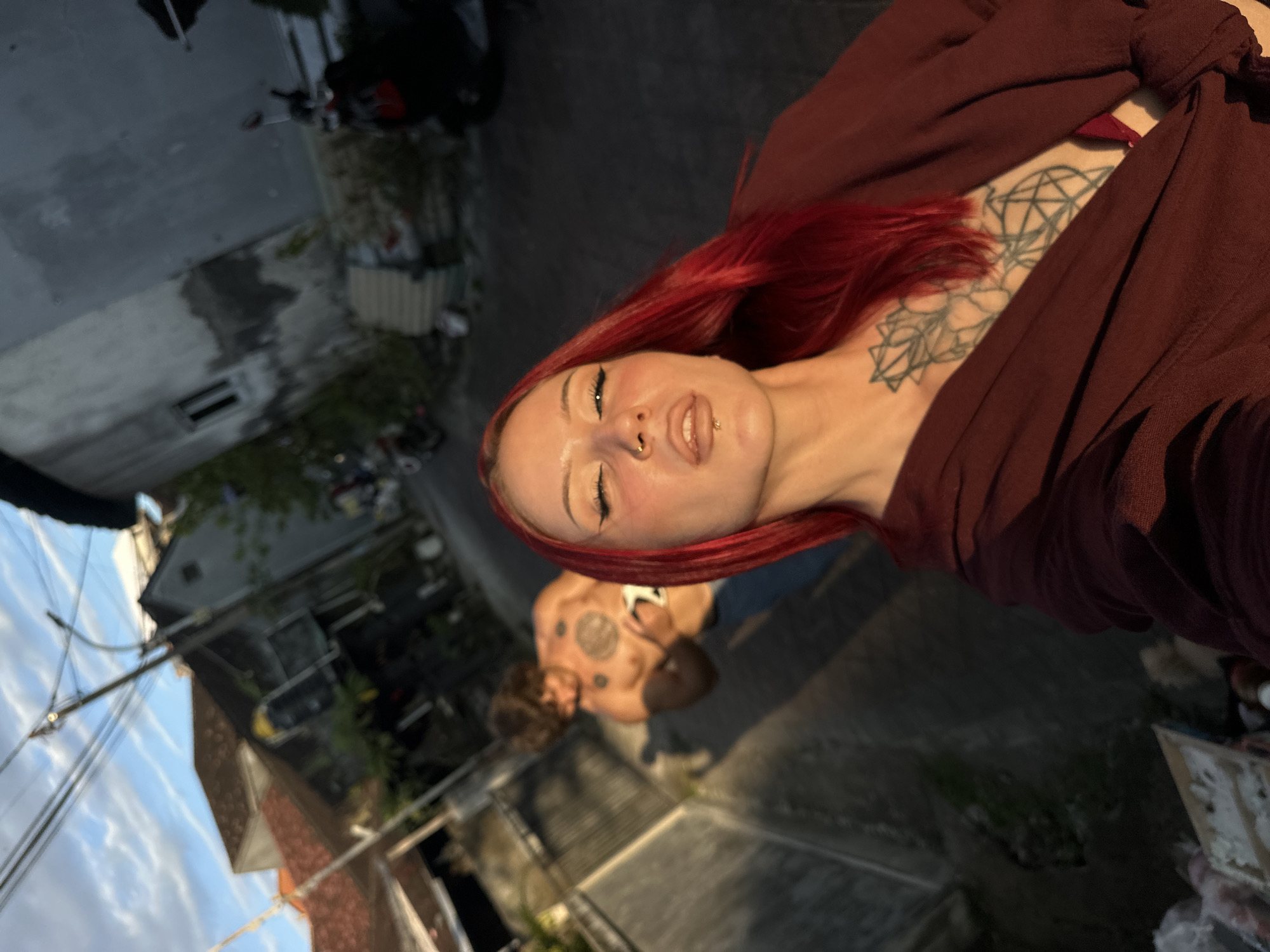 A selfie-style image shows a fair-skinned person with red hair, visible tattoos on their chest, and facial piercings, wearing a dark-colored garment. Another person, shirtless and with visible tattoos, is slightly out of focus in the background. The setting appears to be an outdoor urban environment, with buildings, pavement, and visible sky in the background. The exact location is North Kuta, Indonesia.
