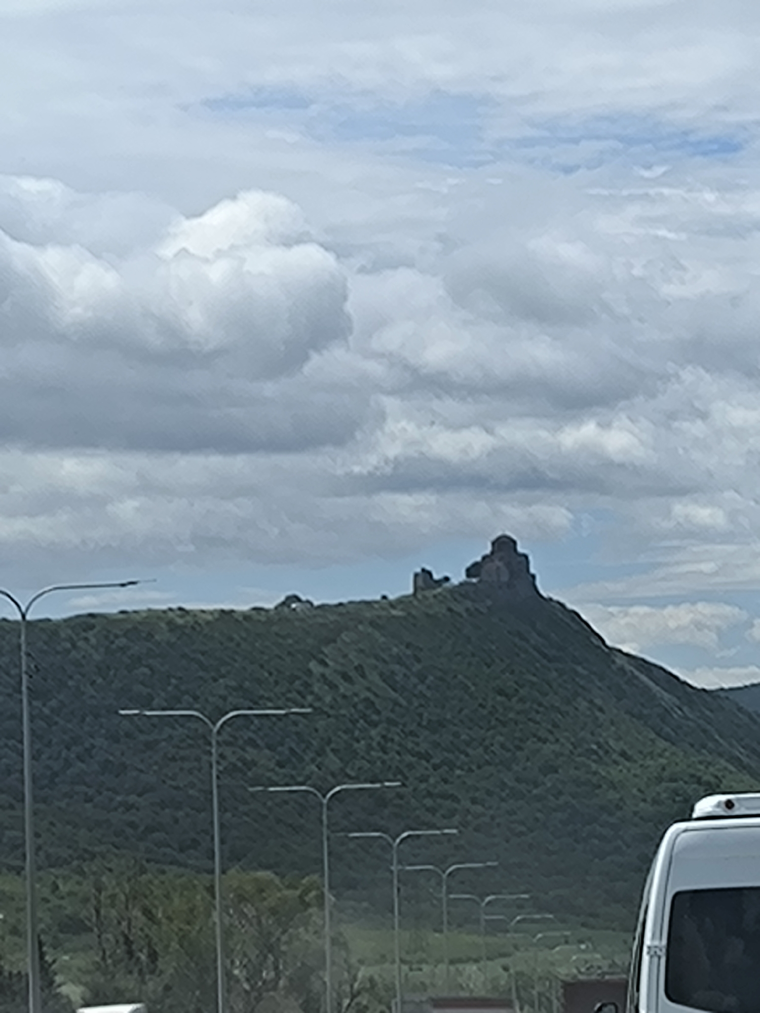 The image shows the Jvari Monastery situated atop a green, tree-covered mountain near the city of Tsitsamuri, Georgia. The sky is filled with grey and white clouds. In the foreground are roadside street lamps and a portion of a white vehicle.
