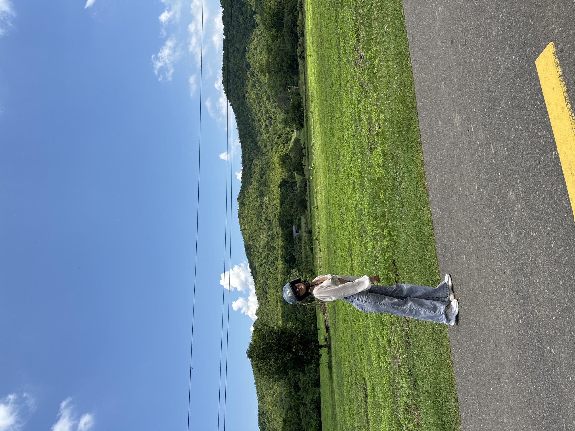 A young woman stands on an asphalt road in Ham Thuan Bac, Vietnam, facing slightly away from the camera and looking over her shoulder. She is wearing a white crop top, blue flared jeans, and a blue helmet. The road has a yellow painted line on its right side. Behind the road is a vibrant green field, followed by a dense forest covering a rolling hillside, and a bright blue sky with scattered white clouds. Three parallel black wires stretch horizontally across the upper portion of the image.