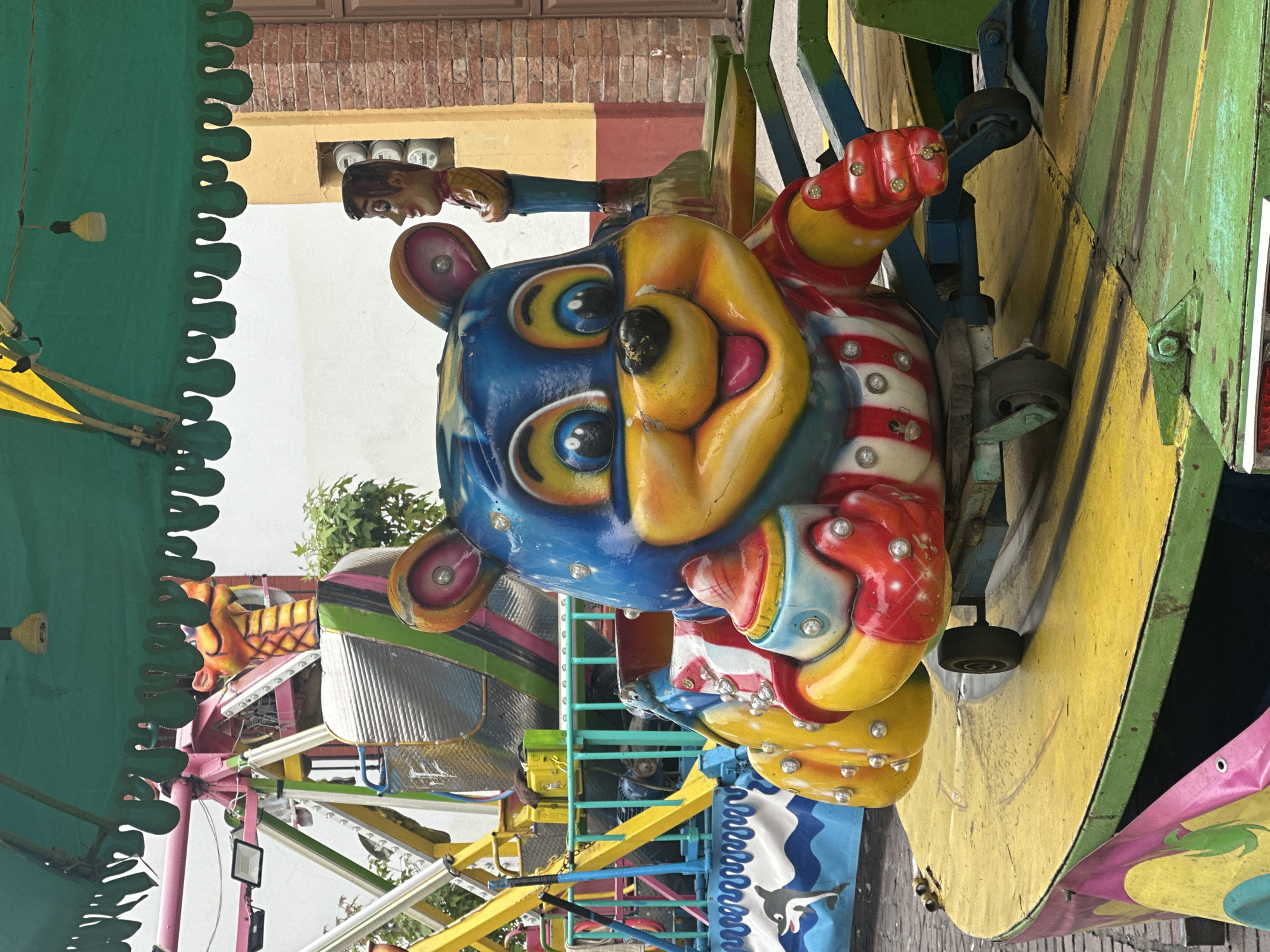 The image depicts a colorful amusement park ride, likely located in San Miguel Zinacantepec, Mexico, although specific urban or geographic markers are absent. The ride features a bear-shaped seat with a blue and yellow face and a striped body. The bear figure is attached to a structure composed of yellow, green, and blue painted metal beams. Another figure of Woody from Toy Story is seen on the upper part of the ride. The ride is under a green scalloped awning. The composition, viewed from below, presents a vibrant scene of a fairground attraction.