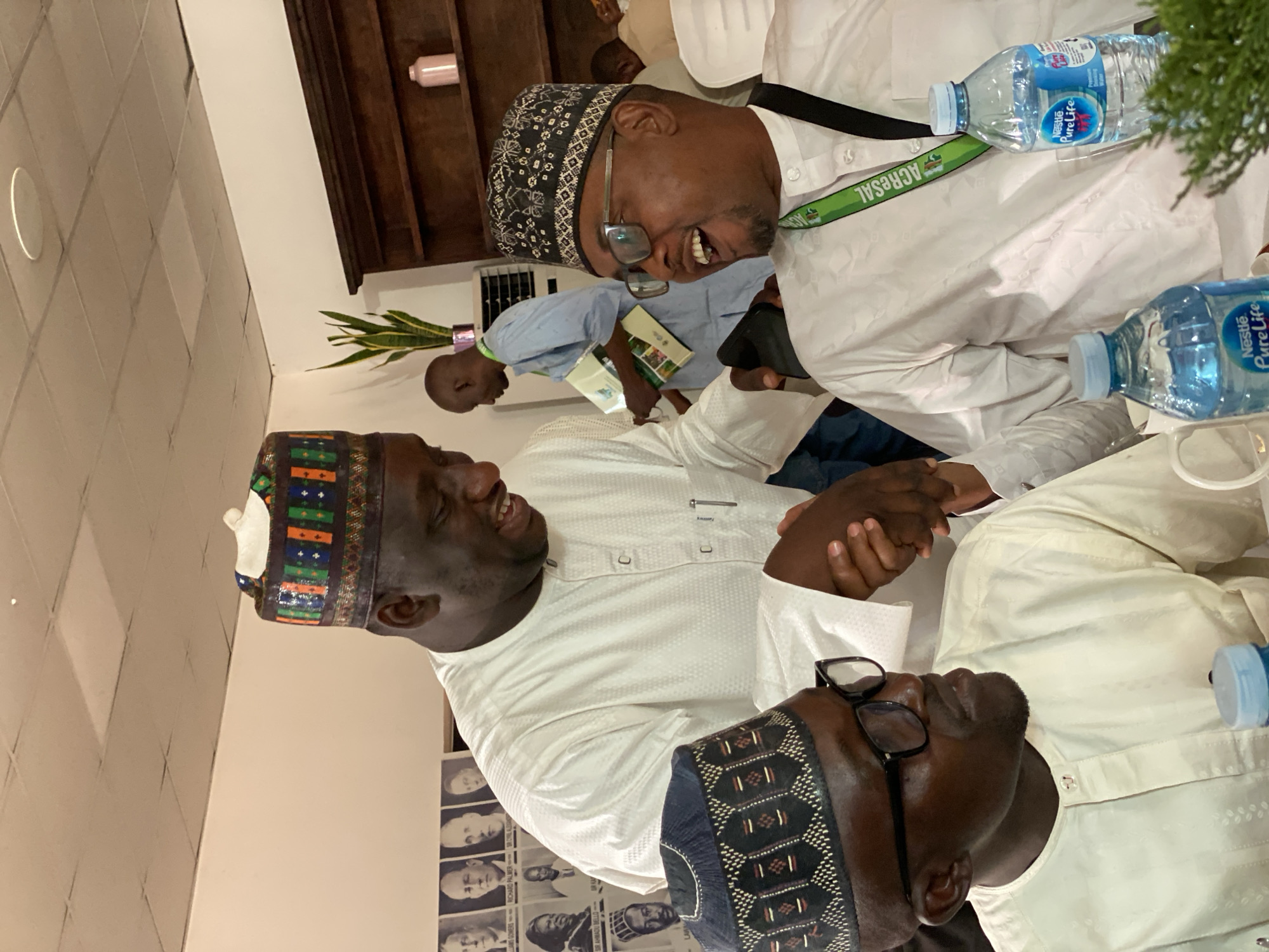The image shows four men in an indoor setting in Kwarau, Nigeria. Three men are wearing traditional caps. The man on the left has a cap with green, orange, and blue embroidery. The man on the right wears a black cap with glasses. The man in the middle is laughing, wearing glasses and a black cap. A lanyard with "ACRESAL" is around his neck. The men are wearing white collared shirts and are shaking hands. Two water bottles are on the table. Behind them is a wall with framed portraits of individuals and a man holding a book.