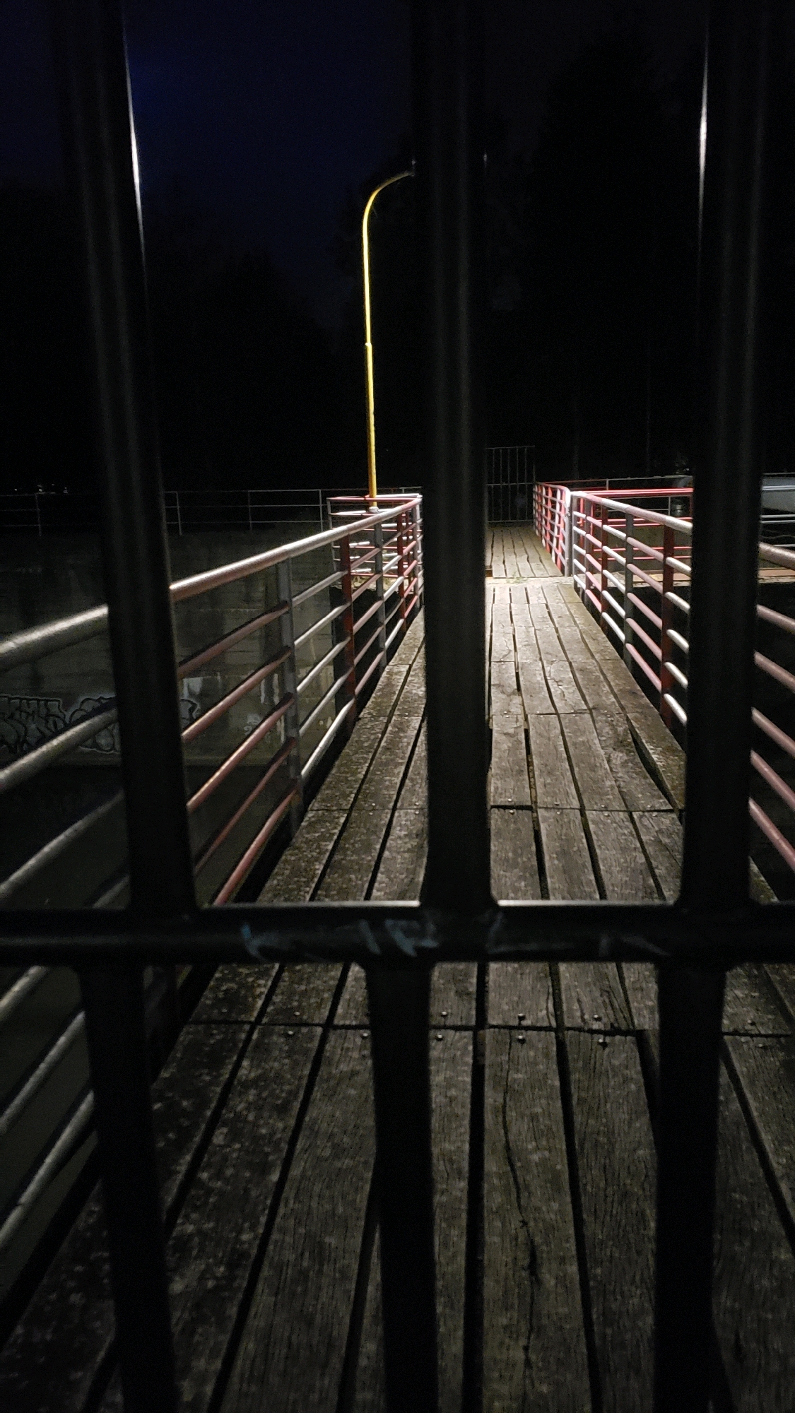 The image depicts a dark, nighttime scene of a wooden boardwalk with metal railings on either side. The boardwalk appears to be elevated, possibly over water or a canal, given the presence of a concrete embankment with graffiti on the left. A single yellow lamppost is visible in the distance, casting a faint light. The foreground is dominated by dark, vertical bars, suggesting the image is taken through a fence or gate. The overall atmosphere is one of quiet and perhaps solitude. Based on the distinctive yellow lampposts and the architectural style of the railings, this scene is likely located at the Sekčovské sídlisko area in Prešov, Slovakia, specifically along the waterfront promenade.