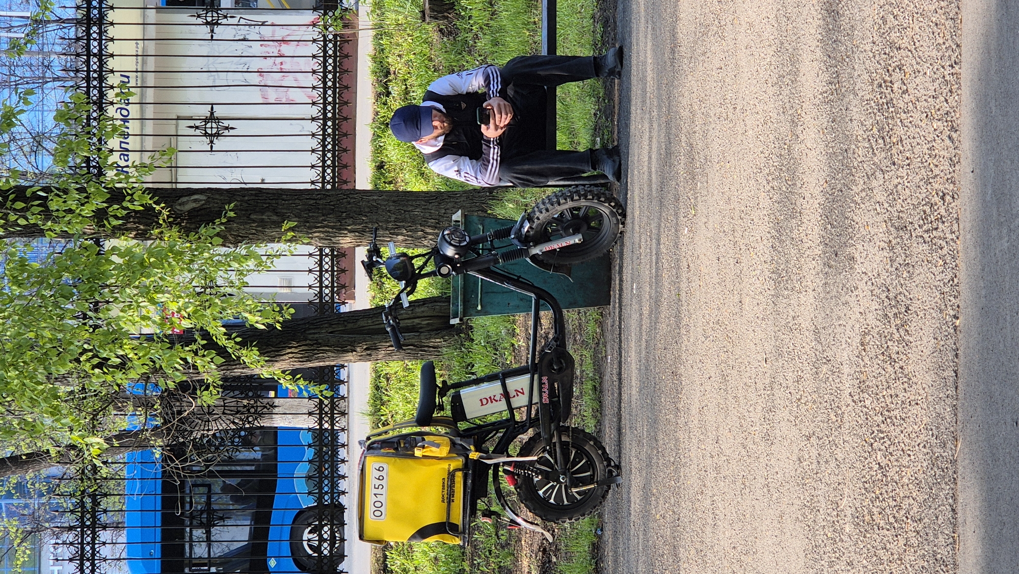 A man sits on a park bench using a smartphone, next to a parked DKALN electric bike. The scene is set in Kemerovo, Russia, as indicated by text on a sign in the background. The bike is yellow and black, with a delivery box attached to the back, displaying the number "001566" and the words "доставка и магазин продуктов" (delivery and grocery store). The man is wearing a dark jacket with white stripes on the sleeves, dark pants, and a blue beanie. A black metal fence and trees are visible in the background, with a blue vehicle parked beyond the fence. The path alongside the bench is paved.