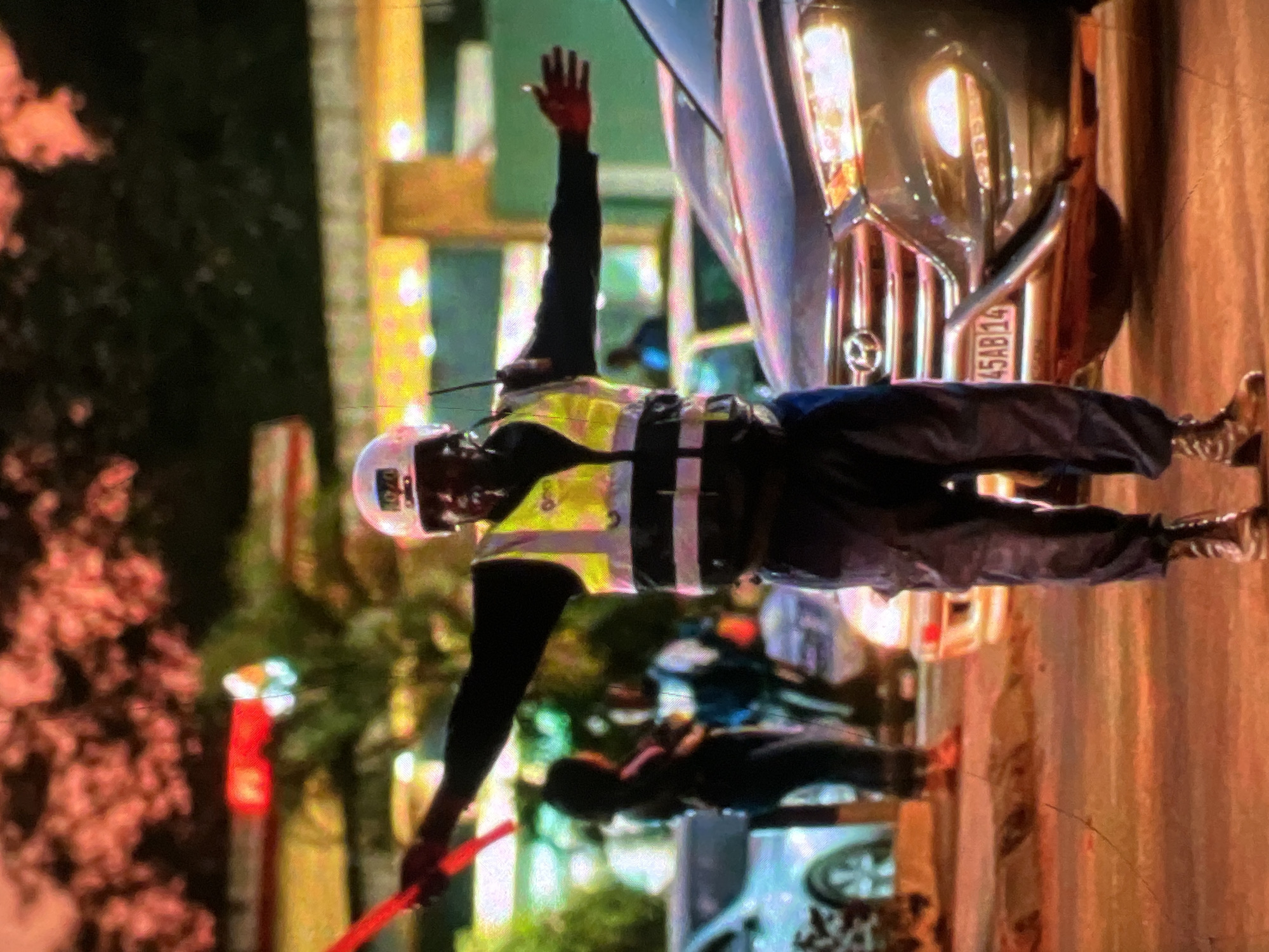 The image captures a traffic director in Lagos, Nigeria, actively managing traffic. He is wearing a reflective yellow vest over a dark long-sleeved shirt and dark pants, with a white helmet on his head. In his hands, he holds two red traffic wands, one extended upwards and to his left, and the other to his right. His arms are outstretched, indicating he is directing vehicles. Behind him, a dark SUV with its headlights on is partially visible, and a license plate reads "45AB14". In the background, other vehicles and pedestrians can be seen, suggesting a busy urban setting. The lighting in the image indicates it is likely dusk or evening, with streetlights and vehicle lights illuminating the scene. The environment appears to be a street or intersection within the city.