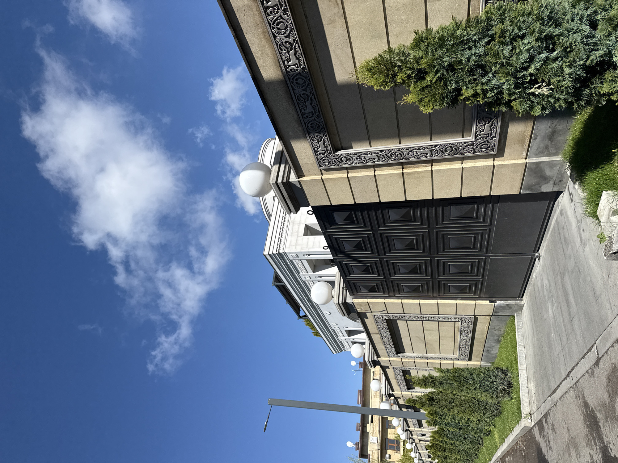 The image shows the exterior of a building in Yerevan, Armenia, under a bright blue sky with scattered clouds. The building has a modern architectural design with decorative stone cladding and black metal gates. Green shrubbery grows along the base of the building and along the sidewalk. A street lamp stands nearby, with other buildings visible further down the street, also lined with lamps.
