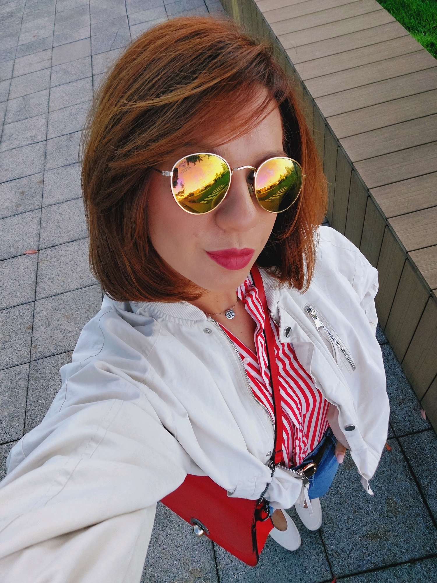 A woman with reddish-brown hair and mirrored sunglasses is taking a selfie outdoors. She is wearing a white bomber jacket over a red and white striped shirt. A red purse is slung over her shoulder. Her lower body is visible, showing blue jeans and white sneakers. The ground is paved with gray tiles, and a wooden structure with green grass is visible in the background. The scene appears to be in a public urban space, possibly a park or plaza, in Sofia, Bulgaria.