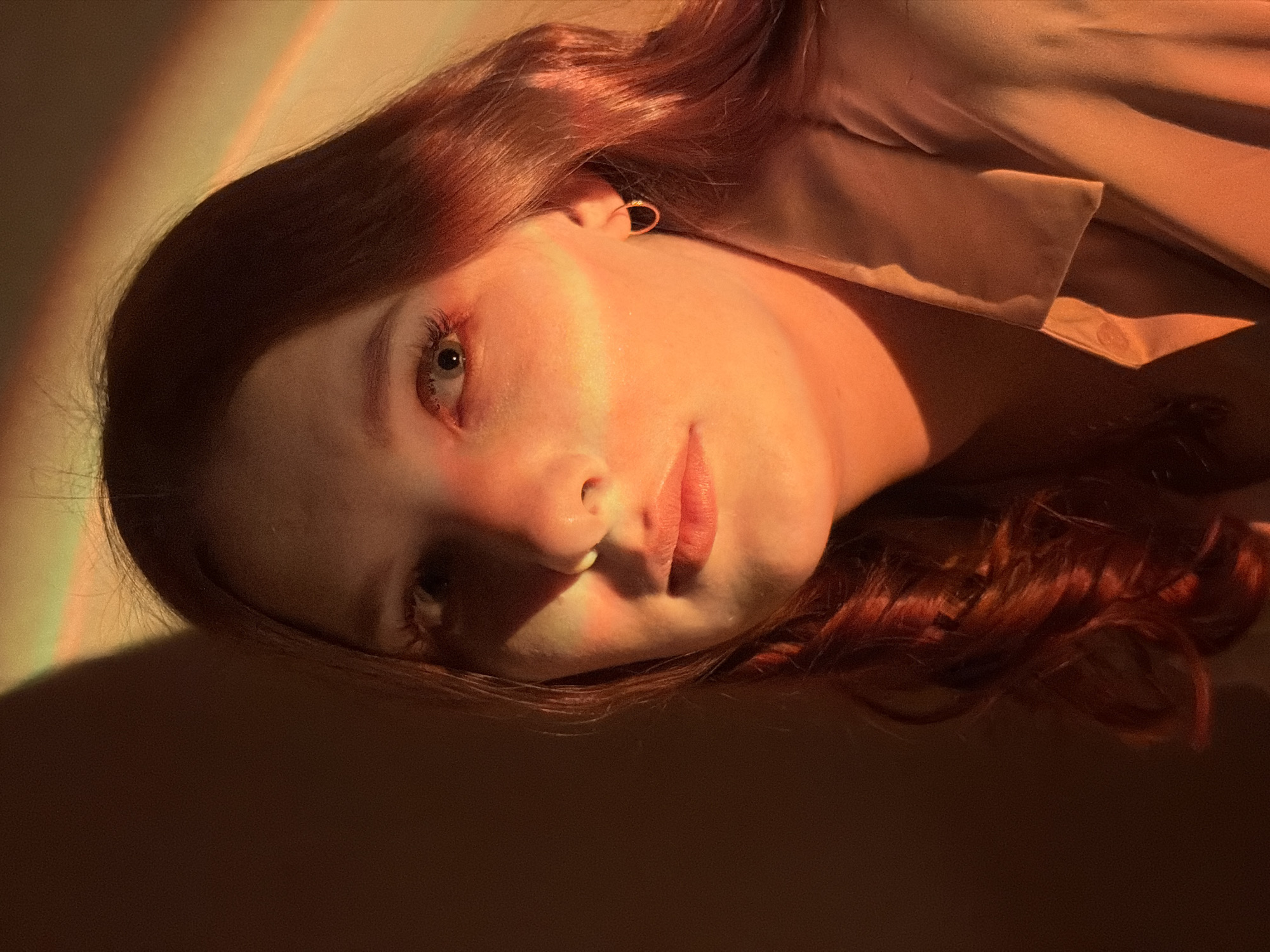 A close-up, warm-toned portrait features a young woman lying down, looking directly towards the viewer. Her head is angled, resting on a flat, plain surface. She has long, wavy, reddish-brown hair that frames her face and spreads around her head. The scene is dramatically lit by a strong, warm light source, possibly sunlight, which casts vibrant orange and red hues across her face, hair, and the background. This light illuminates the left side of her face, highlighting her fair skin, nose, and lips, and creating deep shadows on the right side and behind her. Her lips are slightly parted, and her eyes are open with long lashes. A small, delicate gold-colored hoop earring is visible in her left ear. She appears to be wearing a light-colored, possibly beige or light pink, collared garment, parts of which are visible near her neck and shoulder. The background is simple, predominantly in warm tones, suggesting an indoor setting in Chaikovsky, Russia. The intense, directional lighting gives the image a serene and intimate mood, hinting at either early morning or late afternoon. No text is visible.