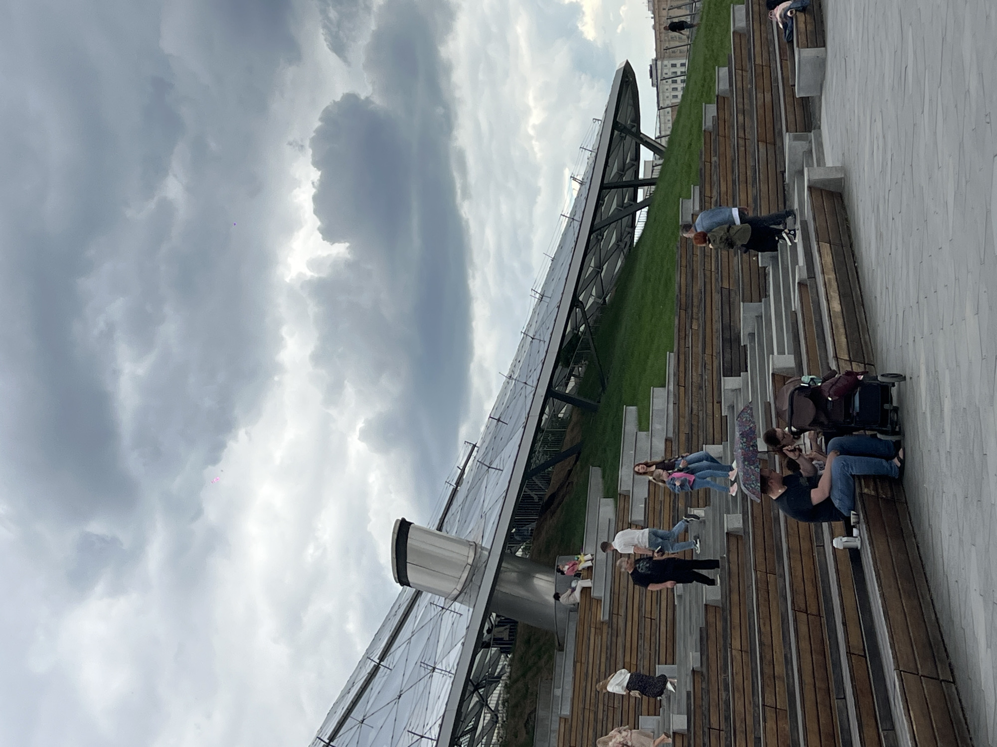 People are relaxing and walking on wooden steps next to a grassy hill and a modern architectural structure in Zaryadye Park, Moscow, Russia. The sky is cloudy.