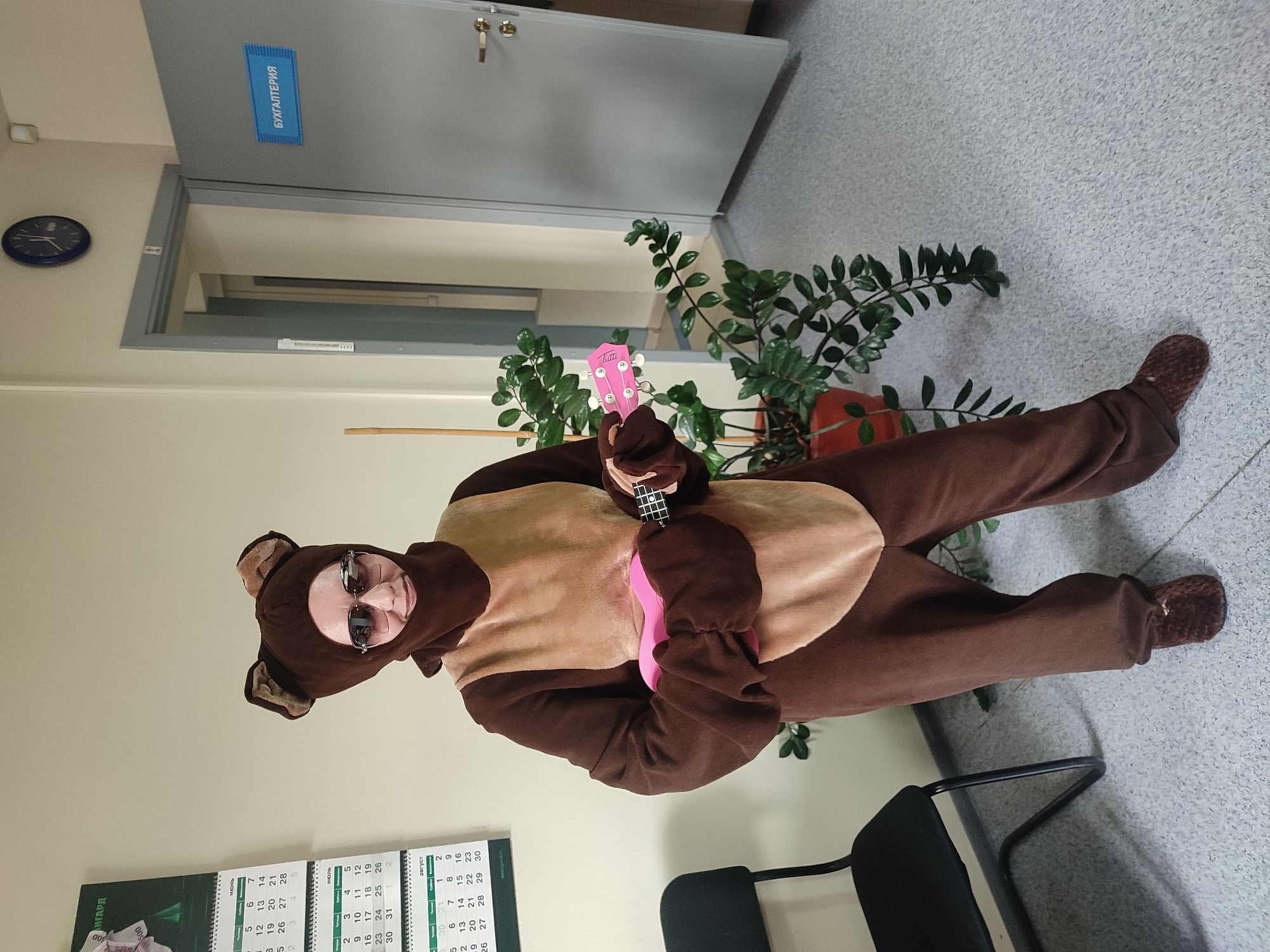 A person wearing a brown bear costume, complete with ears and a light brown chest patch, stands in an indoor hallway. The person is also wearing large sunglasses. They are holding a pink ukulele in their right hand and a black object, possibly a phone, in their left hand. To the left of the person is a black chair, and to their right is a potted green plant. Behind the plant is a grey door with a blue sign that reads "БУХГАЛТЕРИЯ" (Accounting). A calendar is visible on the wall to the left. The floor is a light grey speckled material. The scene appears to be in an office or institutional setting.