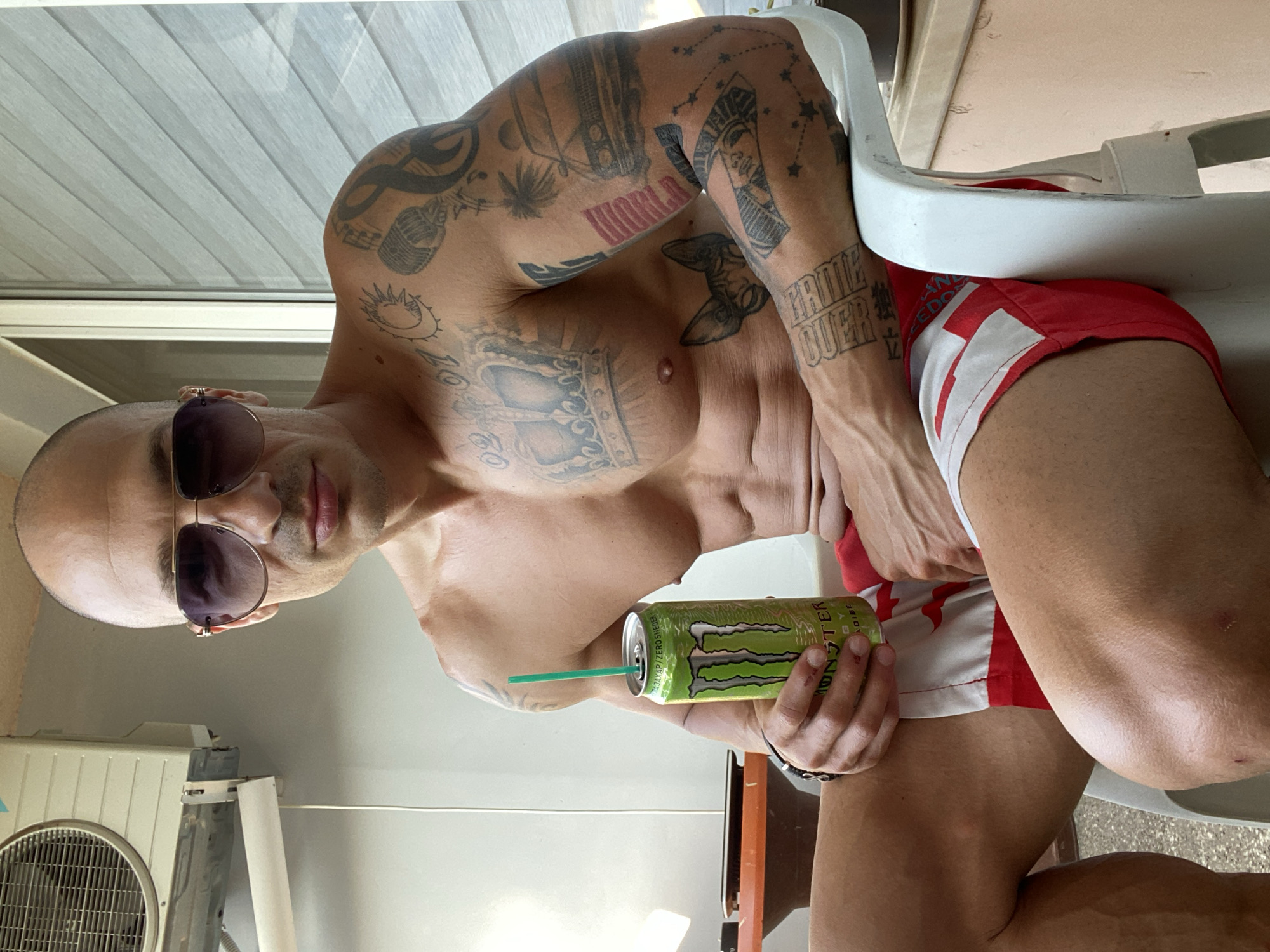 A bald, tattooed man wearing sunglasses and red and white shorts sits on a white plastic chair outdoors. He holds a green can of Monster Energy drink with a straw. The man is shirtless and displays his muscular physique. In the background, an air conditioning unit is visible on a light-colored wall, and blinds are drawn on a window. The environment appears to be a balcony or patio area.