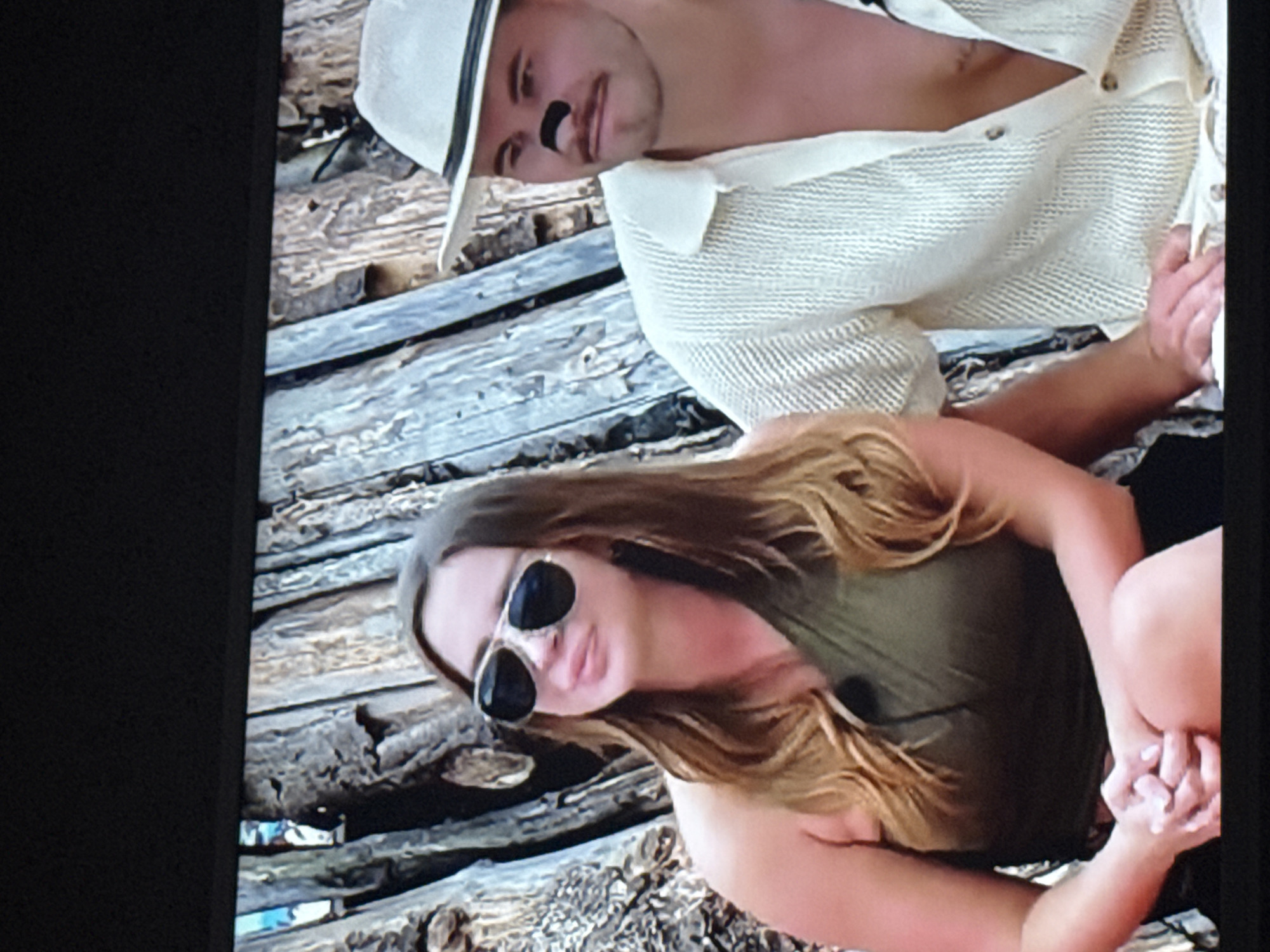 A man and a woman are sitting on a wooden structure. The man is wearing a white hat and a white button-down shirt. He has a dark shape under his nose. The woman is wearing sunglasses and a green top. They are on a structure made of rough logs or planks.
The location cannot be determined from the image.
