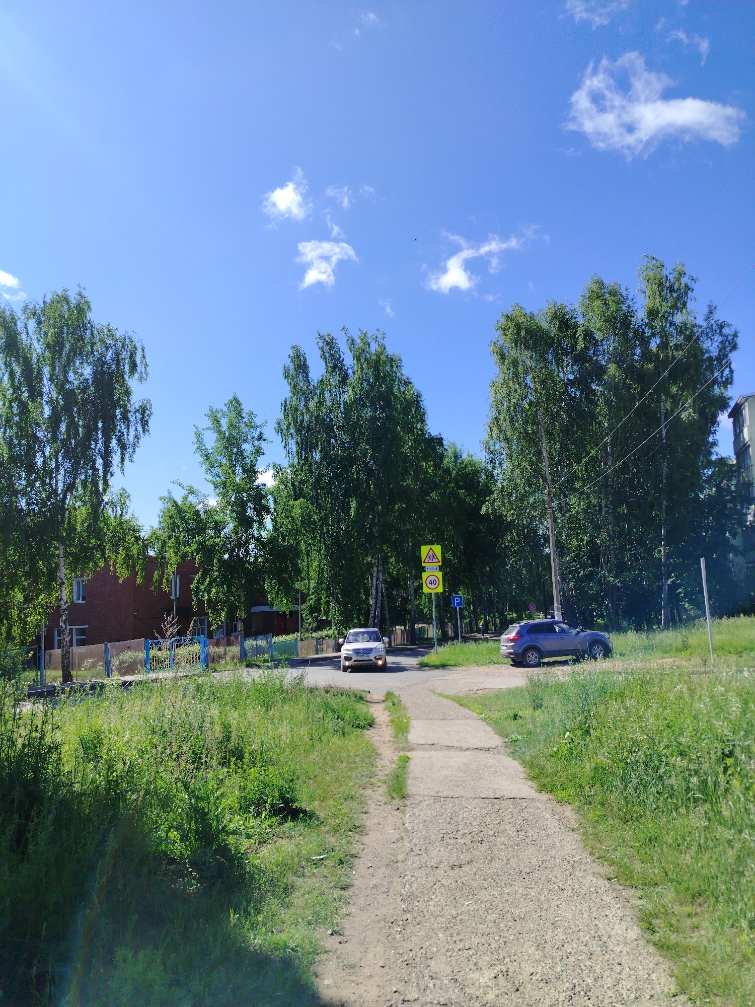 The image depicts a sunny day in Noviy, Russia, with a clear blue sky dotted with a few white clouds. A dirt path winds through lush green grass, leading towards a paved road. On the road, two cars are visible: a white sedan and a dark-colored SUV. Beside the road, a yellow triangular sign with children walking is posted, along with a circular sign displaying the number "40," indicating a speed limit. A blue metal fence runs along the property line of a red brick building with several windows. Tall, leafy green trees are abundant throughout the scene, their branches reaching towards the sky. The environment suggests a residential area with a mix of natural and developed elements.