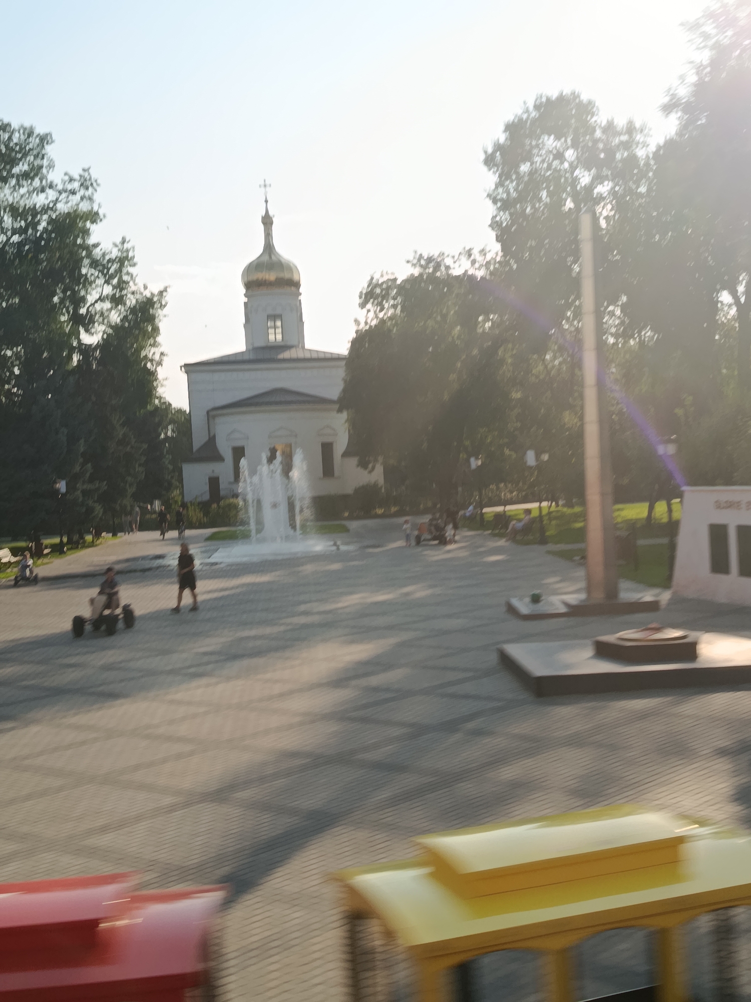 The image captures a bright, sunny day in what appears to be a city park or public square. In the background, a white building with a prominent gold-domed cupola, characteristic of an Orthodox church, stands against a clear sky. In front of the church, a multi-tiered water fountain is active, with streams of water arcing upwards.
The foreground is paved with rectangular grey tiles arranged in a grid pattern. Several people are visible enjoying the outdoor space. Two individuals are operating pedal go-karts. One person is walking towards the viewer, and another appears to be seated on a go-kart. Further back, a group of people are gathered near the fountain and on benches, suggesting a relaxed atmosphere. Trees with lush green foliage line the perimeter of the square.
In the mid-ground, a tall, slender monument or column stands on a raised platform. To the right of this monument, a light-colored structure with dark rectangular panels is visible, possibly a memorial. In the immediate foreground, the blurred forms of red and yellow vehicles, perhaps part of a tourist attraction or a small amusement ride, are present, indicating movement and a sense of dynamism.
The overall impression is one of a lively public space, possibly a town center, where people are engaging in recreational activities amidst a backdrop of architectural and memorial elements. Based on the provided context, this scene is located in Cahul, Moldova.