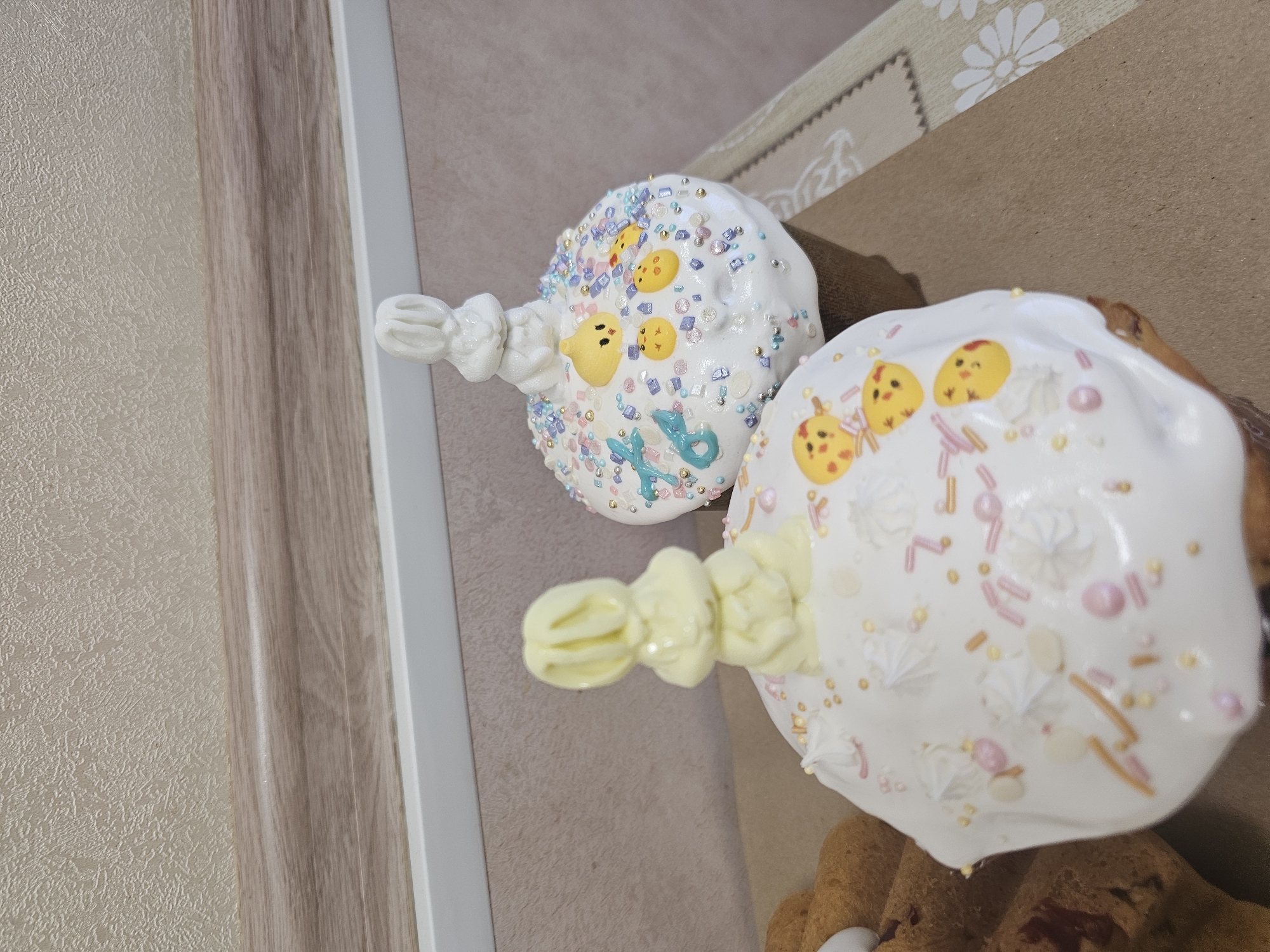 Two Easter cakes are shown in the image. They are decorated with white icing, sprinkles, and small yellow chick-shaped decorations. One cake has a white icing decoration extending outward, while the other has a yellow icing decoration. The cakes are placed on a brown surface. A wall with a light brown and beige design is visible in the background. The image was probably taken indoors. No specific details indicate the location is in Moscow, Russia.