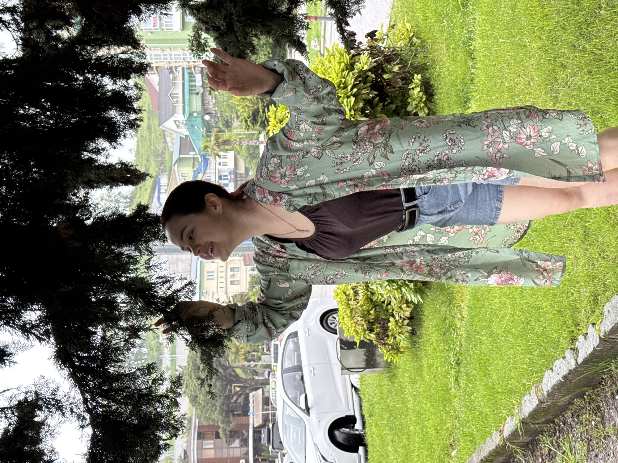 A woman stands on a street in Sochi, Russia. She wears a brown top, denim shorts, and a long, light green floral print jacket. She is touching the branches of a coniferous tree. Behind her are parked cars, green foliage, and multi-story buildings. The ground is covered with green grass.
