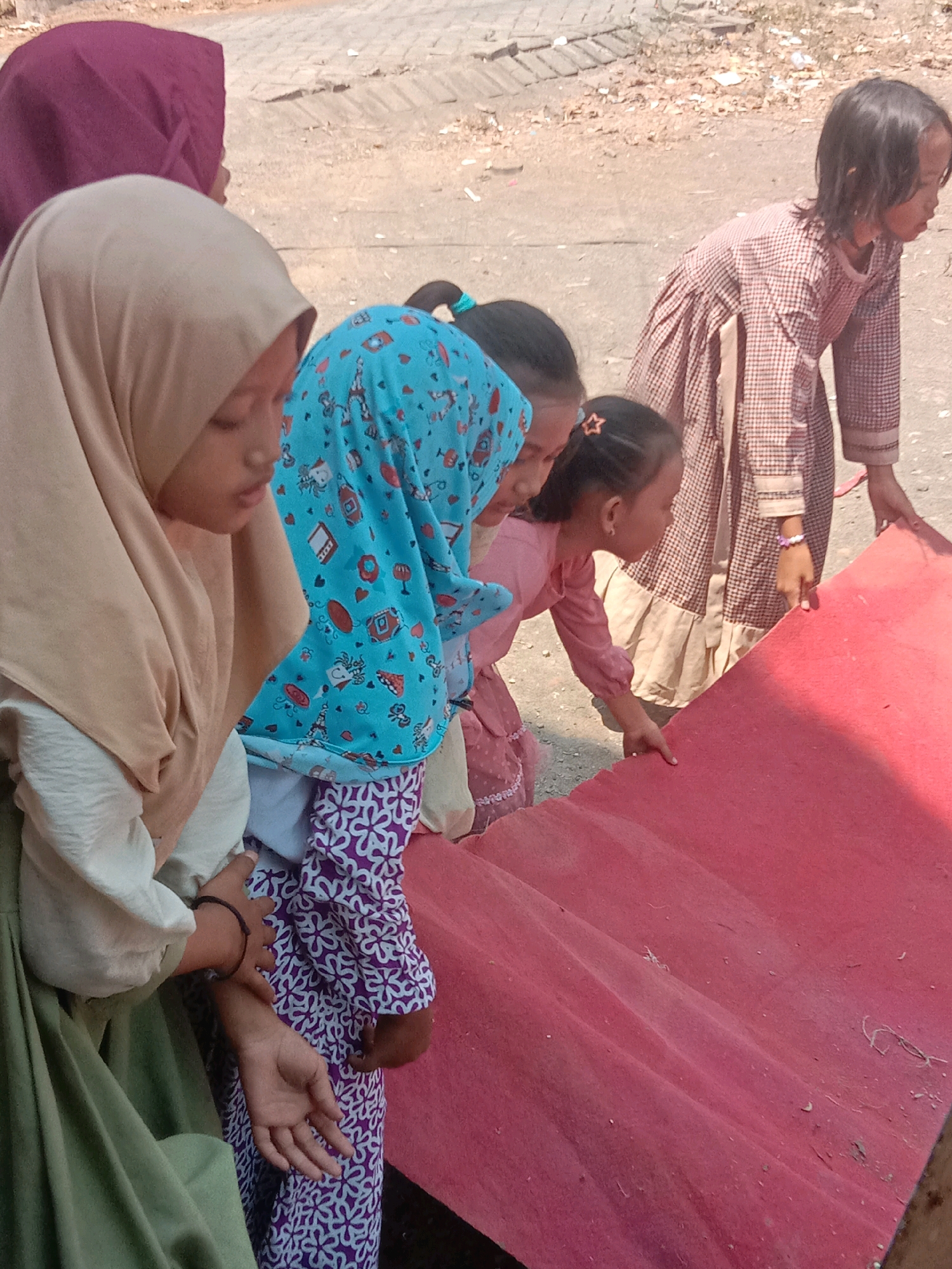 The image depicts five young girls outdoors, in what appears to be a residential area in Karang Anyar, Indonesia, likely on a dusty, paved surface with sparse vegetation. The girls are positioned around a large, folded piece of red material, possibly a rug or a tarp, which dominates the foreground and extends diagonally across the frame.
From left to right, the first girl is wearing a light green dress and a tan hijab, with her head bowed and her hands clasped. Next to her is a girl in a blue hijab adorned with a playful pattern of cartoon characters and hearts. She is looking downwards. To her right, a girl in a pink dress with puff sleeves and a star-shaped hair clip is looking towards the red material and appears to be touching it. Another girl, in a brown and white checkered dress with a ruffled hem, is also looking at the red material, her hands are near it. The fifth girl, on the far right, is mostly out of frame but her hands are visible on the red material.
The environment suggests a warm, sunny day. The background shows a dirt path with some scattered debris and evidence of construction or uneven paving. The overall scene conveys a sense of casual activity among the children.
