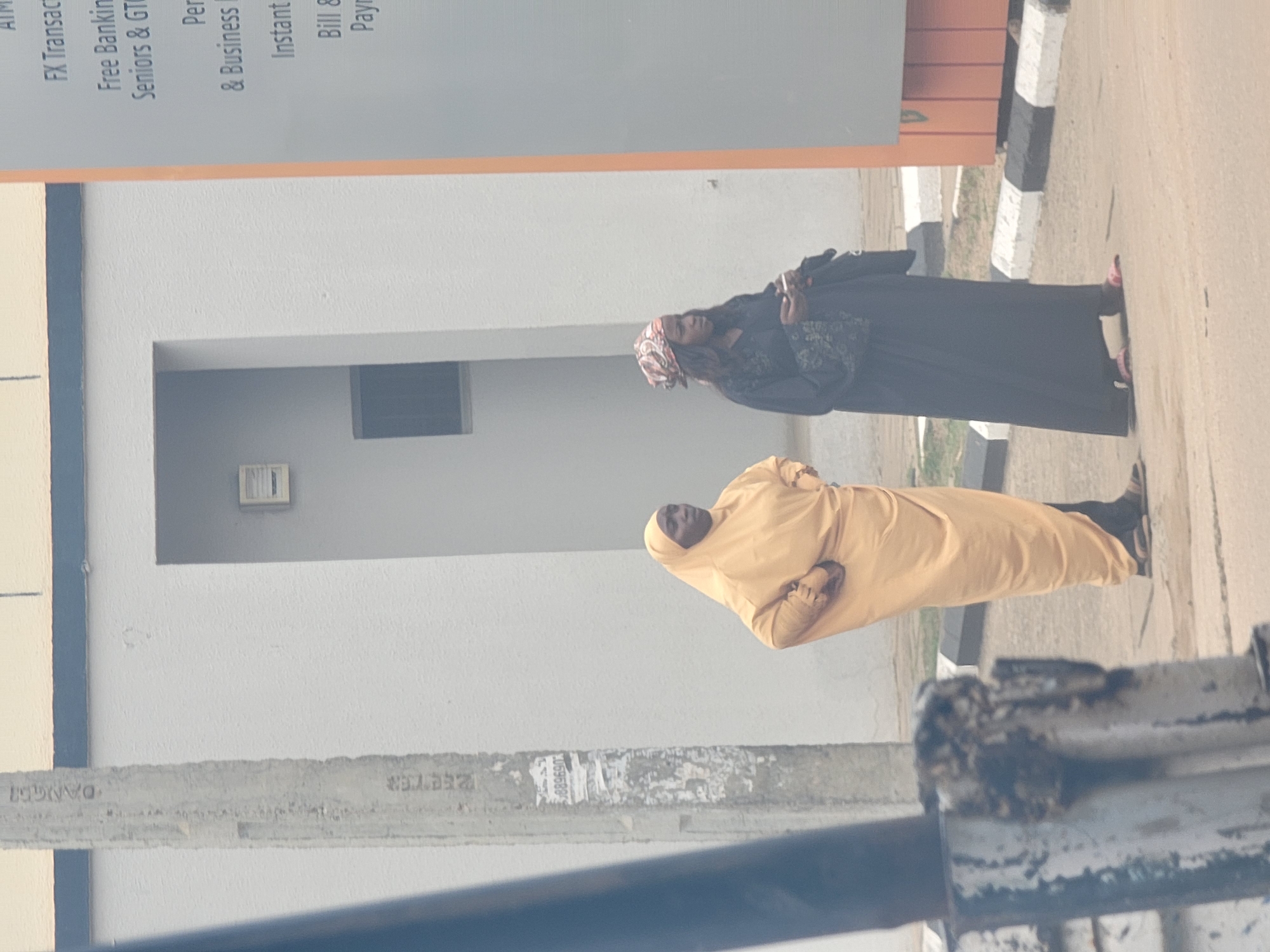 The image shows two women standing on a sidewalk in what appears to be Maiburiji, Nigeria. One woman is wearing a light yellow head-to-toe covering, while the other wears a dark colored garment and a patterned headscarf. A building with text promoting financial services is in the background. The text includes "FX Transac", "Free Banking", "Seniors & GT", "Per & Business", "Instant", and "Bill & Payn". A metal barrier is visible in the foreground. The environment appears urban, with typical street features like sidewalks and painted curb markings.