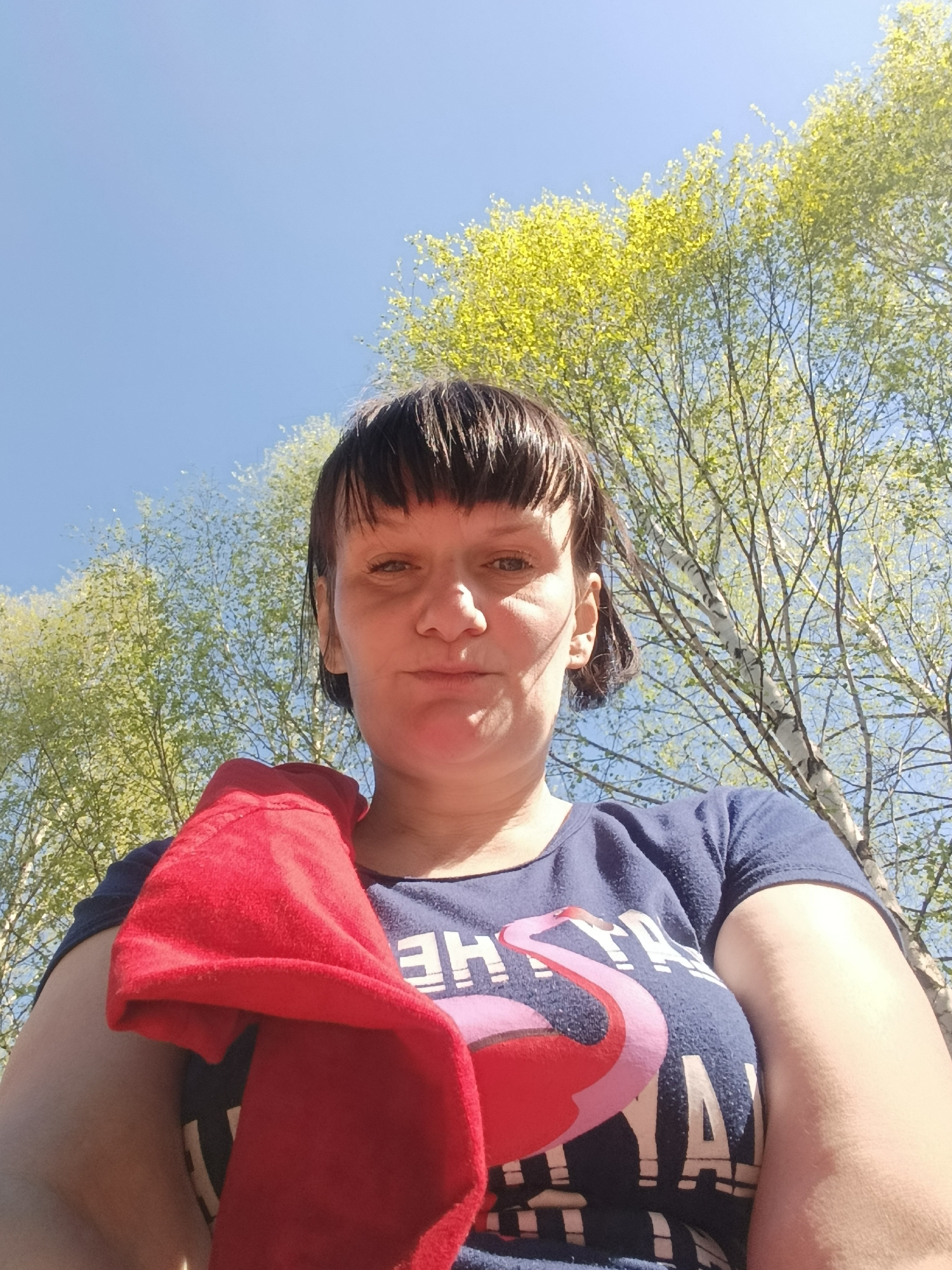 A woman with short, dark hair and a red cloth over her left shoulder is looking directly at the camera from a low angle. She is wearing a navy blue t-shirt with a flamingo design. Behind her are trees with light green leaves against a blue sky. The location is Фоки, Russia.