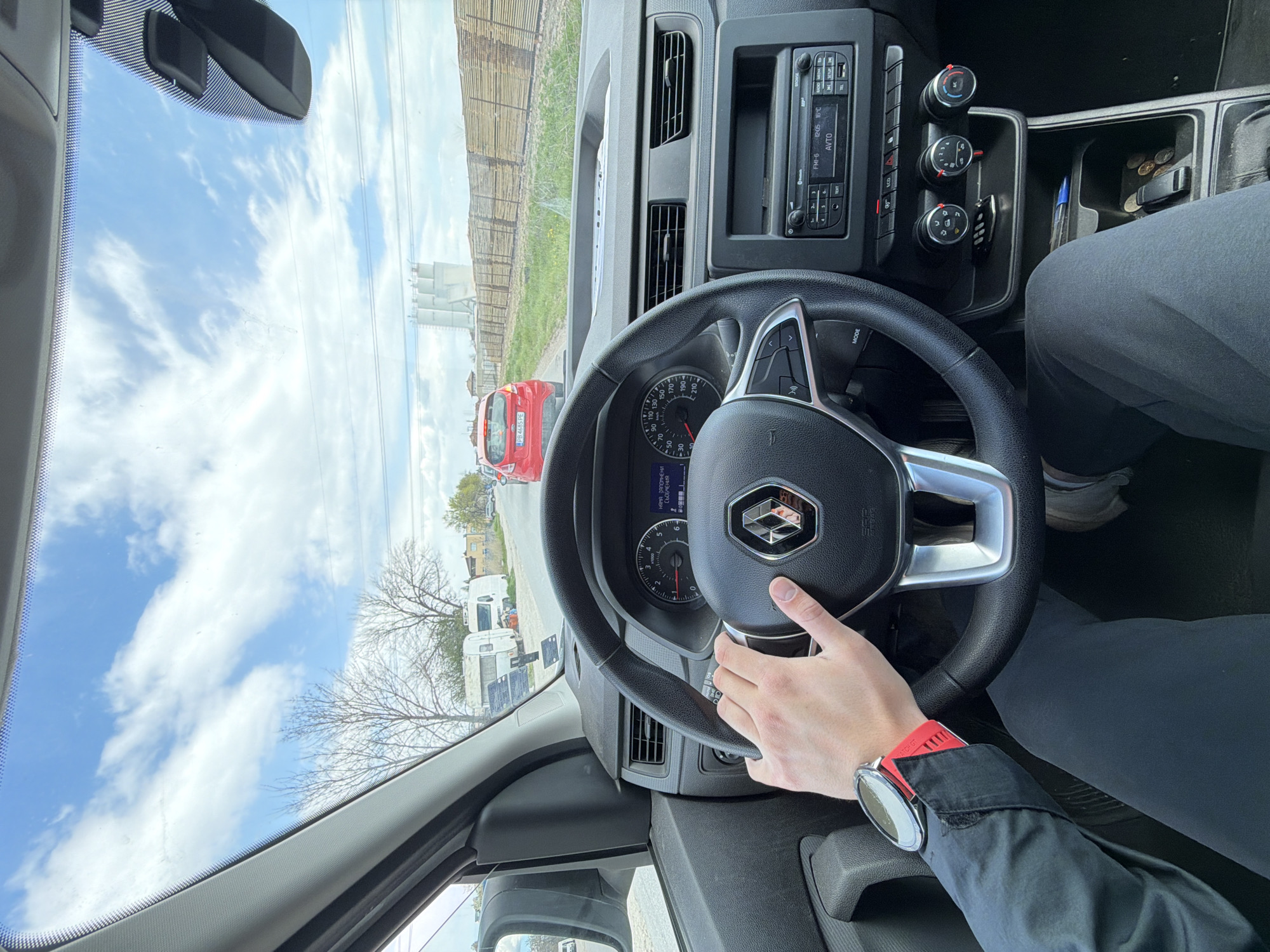The image depicts an interior view from the driver's seat of a vehicle, likely a Renault based on the steering wheel emblem, with the driver's hand visible holding the wheel. A watch with a red band is on the driver's wrist. The vehicle appears to be stationary, as cars and trailers are visible through the windshield along with a red car with the license plate "PB 4685 PE". The location appears to be in the city of Plovdiv, Bulgaria, based on the car's license plate. The sky is partially cloudy. The vehicle's dashboard and radio are visible, and the radio display shows a temperature of 16°C. The center console contains various items including a pen, car key, coins, and other items.
