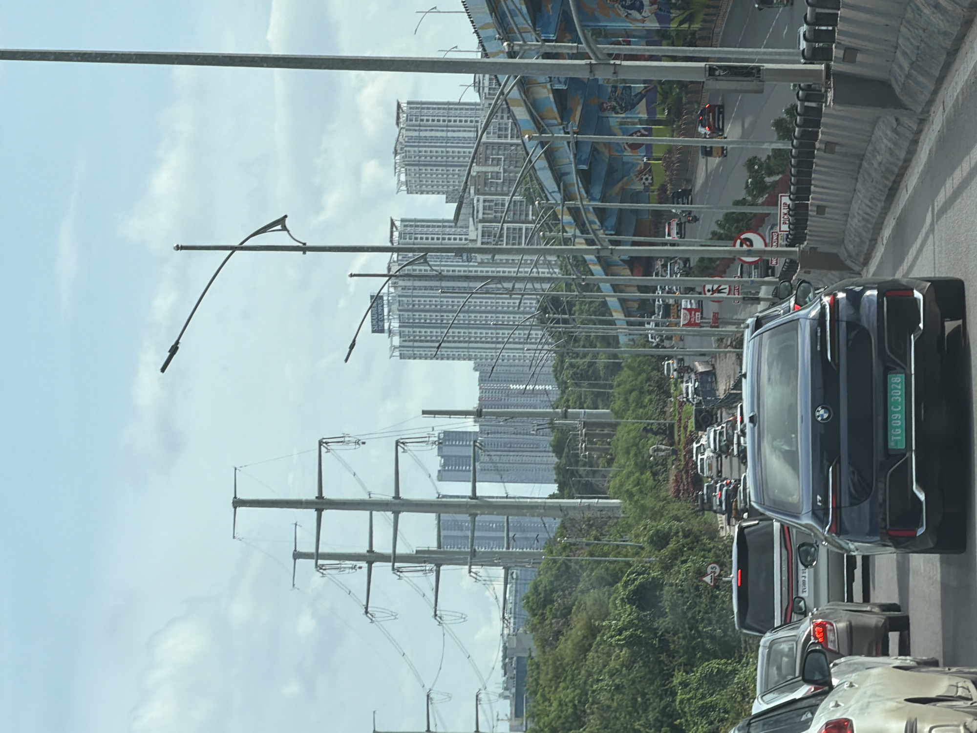 The image depicts a traffic jam on an elevated road in Hyderabad, India, likely during the daytime given the bright sky. In the foreground, a dark blue BMW sedan with license plate TG09C3028 is visible, followed by other vehicles including a white SUV and several cars. Power lines and tall electrical towers dominate the left side of the frame, suggesting proximity to significant infrastructure. In the background, numerous high-rise apartment buildings, including those labeled "SUMADHRUTHI THE OLYMPUS," indicate an urban setting. The road is lined with concrete barriers and poles supporting streetlights. The elevated road itself is adorned with colorful murals featuring depictions of soccer players. Lush green trees and foliage are visible on the embankment beside the road. The sky is partly cloudy.