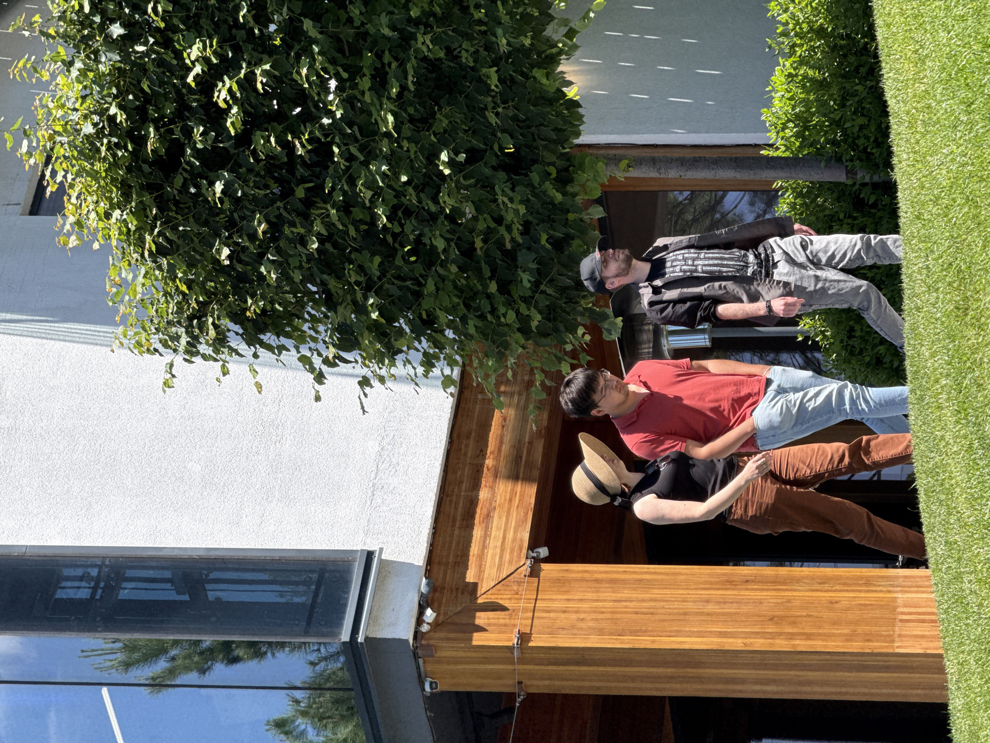 Three people walk on a grassy area in front of a modern building with a large glass window and white stucco walls. A large green tree is situated to the right of the building. The person on the left is a woman wearing a straw hat, a black tank top, and brown pants. The middle person is a man wearing a red polo shirt and blue jeans. The person on the right is a man wearing a black baseball cap, a patterned black and gray shirt, and gray pants. The building has wooden accents around the entrance.