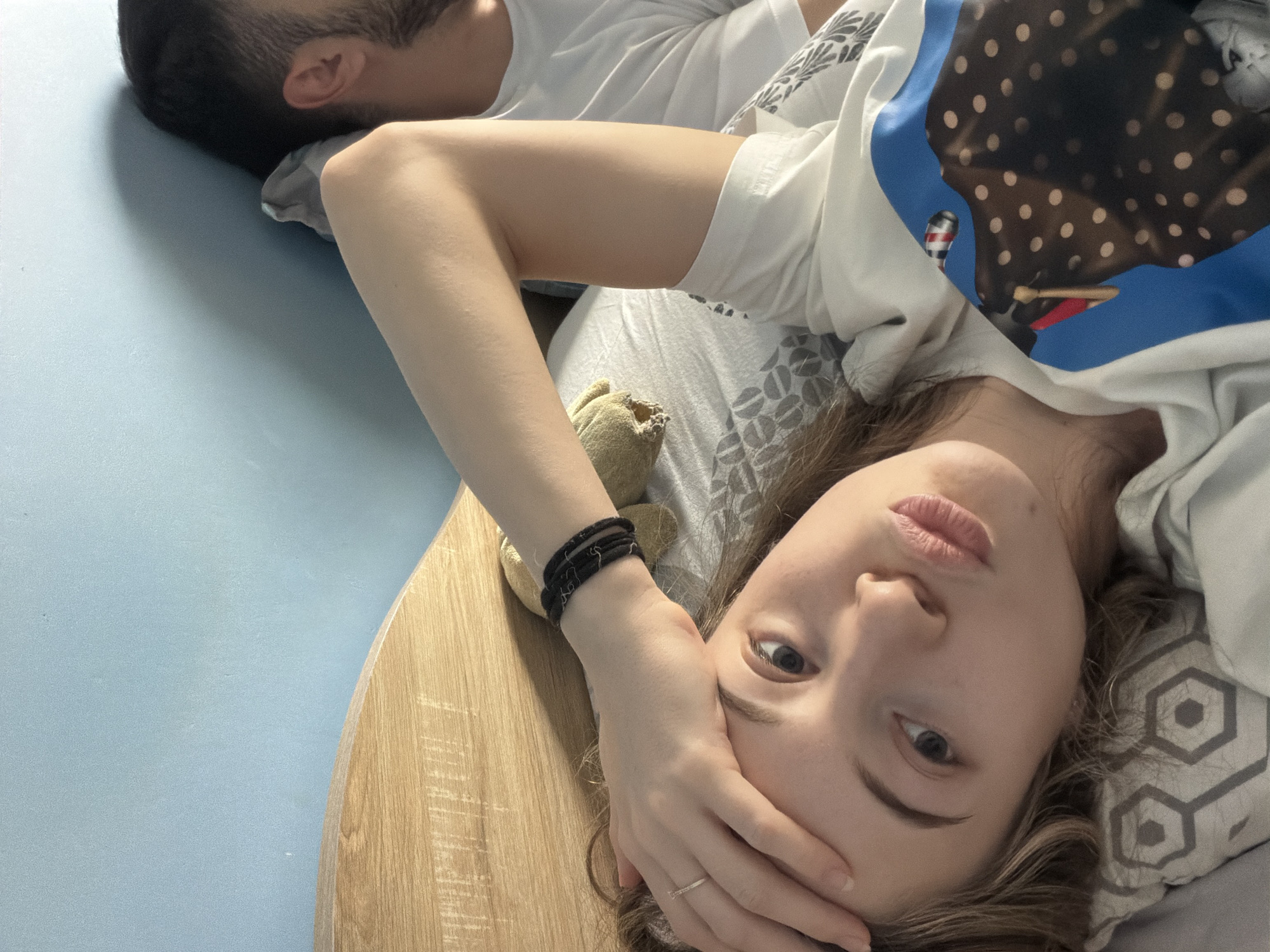 A young fair-skinned woman is lying on a bed, her head resting on a patterned pillow. She is facing the camera, her hand covering part of her forehead. She is wearing a ring on her finger and multiple black bands on her wrist. To her left, a small, stuffed animal toy lies near the edge of the bed. A man is lying next to her, out of focus, wearing a white t-shirt. The bed has a light blue headboard, and the sheets are patterned in grey and white. The location is unspecified and can't be identified as Plovdiv, Bulgaria from the image alone.