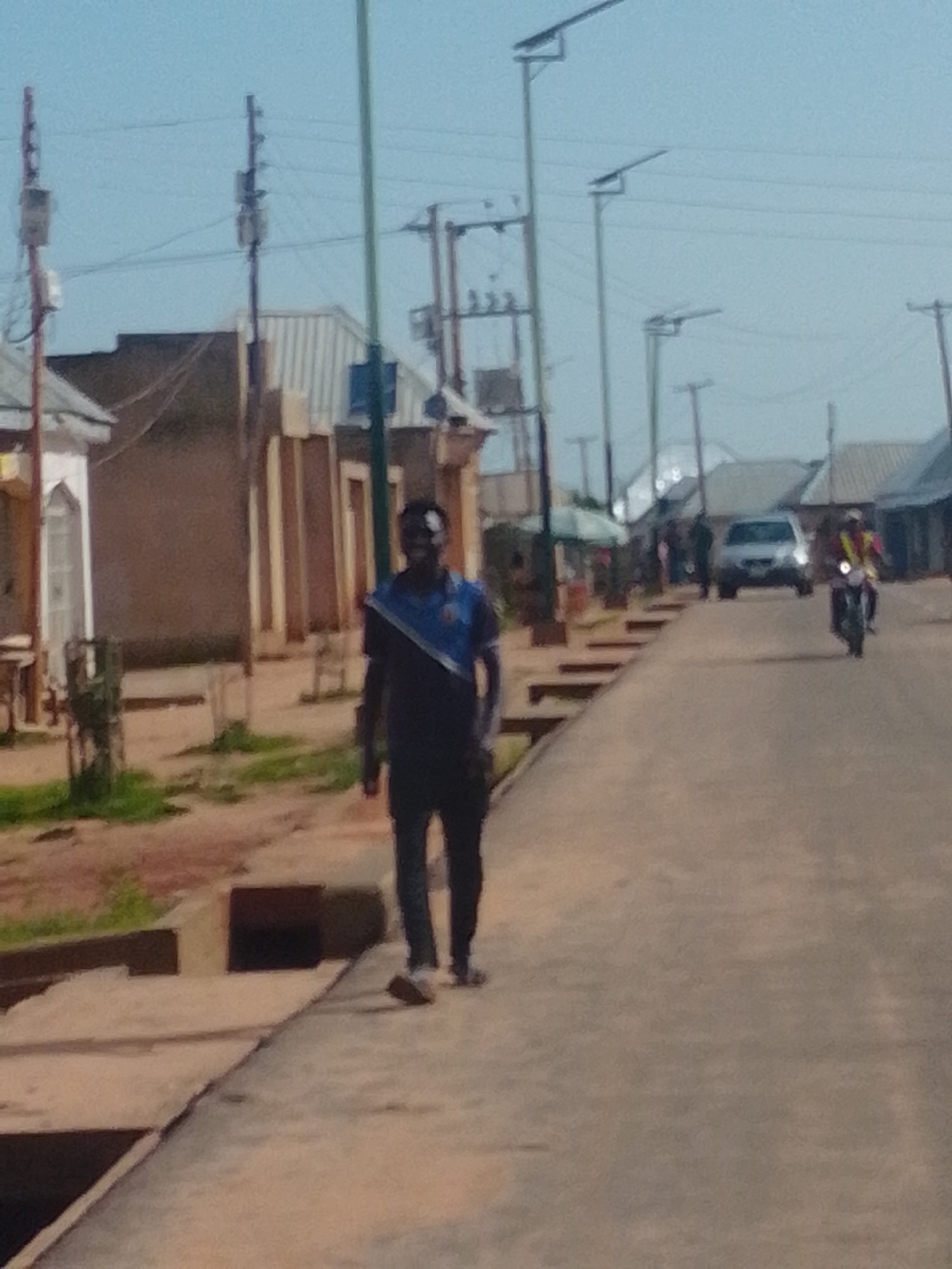 A medium shot captures a man walking on a paved street in what appears to be Gombe, Nigeria. He is wearing a dark blue t-shirt with a diagonal blue and white stripe across the chest, and dark pants. The man is walking towards the camera, and his face is partially visible, looking towards the viewer. The street is lined with buildings on the left, with a mix of structures including a white building with arched windows and another darker building with vertical paneling. Utility poles and streetlights are visible along the street. On the right side of the street, a silver car is parked, and a person on a motorcycle, wearing a yellow vest, is riding past. The ground is a mix of paved areas and dirt with some patches of grass. The sky is a light blue, suggesting daytime.