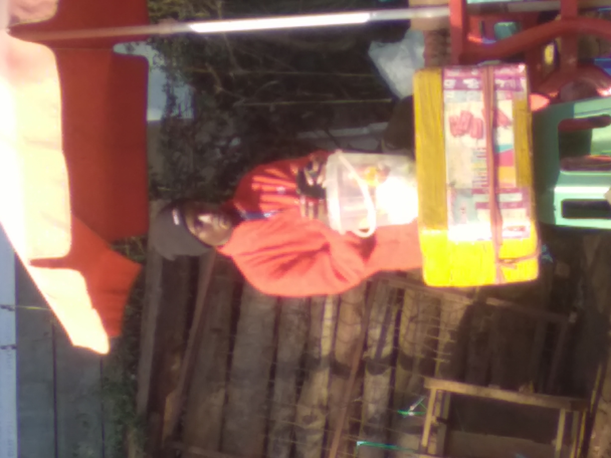 In Nyeri, Kenya, a person wearing a red jacket and a black beanie is seen standing behind a stall. The stall features an orange umbrella overhead and a yellow box filled with items, likely for sale, with colorful packaging visible. In front of the stall, a clear plastic bucket is present. The background consists of wooden structures and lush greenery, suggesting an outdoor market or street vendor setting.