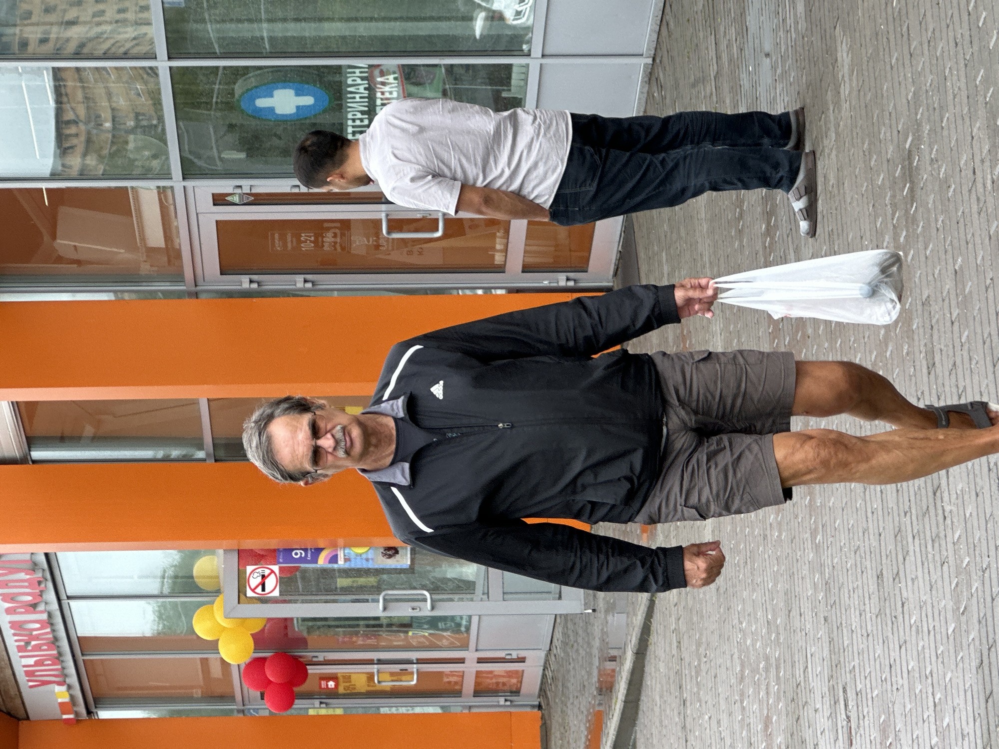 A man walks towards the camera on a cobblestone sidewalk outside a building with an orange facade and large glass windows. He is wearing a black zip-up jacket with white accents on the collar and shoulders, gray cargo shorts, and dark sandals. In his right hand, he holds a white plastic shopping bag. He has gray hair and a mustache and wears glasses. Behind him, another man stands with his hands in his pockets, wearing a light-colored short-sleeved shirt and dark pants. Balloons in red and yellow are visible through the windows on the left, and a "no smoking" sign is also present. The building appears to be a commercial establishment. The image suggests a street scene in Podolsk, Russia, with the Cyrillic lettering on the building indicating a Russian location.