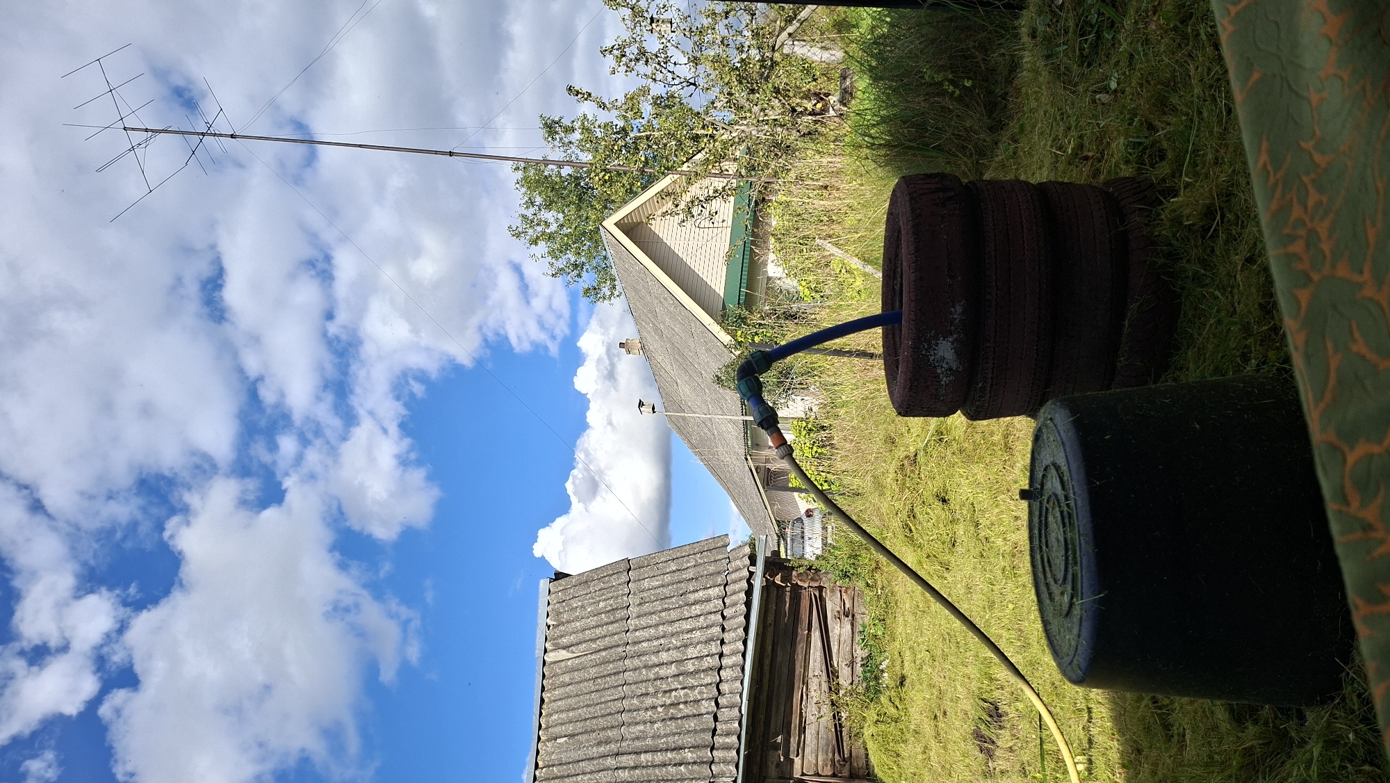 In an outdoor scene located in Svatkavski Self-Governing Council, Belarus, a bright blue sky with scattered white clouds dominates the upper portion of the image. A tall antenna, consisting of horizontal and vertical elements, is erected on a pole in the middle ground. To the left, a rustic wooden building with a corrugated roof is visible, with another house featuring a grey, corrugated roof and green trim in the background. In the foreground, a large black plastic tub with a circular pattern on its lid sits on green, mown grass. To the right of the tub, a stack of four old tires, some with visible wear and paint, are arranged vertically. A yellow hose snakes from the left, connecting to a blue hose that extends from the yellow hose into the top tire of the stack. The surrounding area is covered in grass, with some overgrown vegetation and a chain-link fence visible in the far right. The overall scene suggests a rural or semi-rural setting, possibly a backyard or garden area.