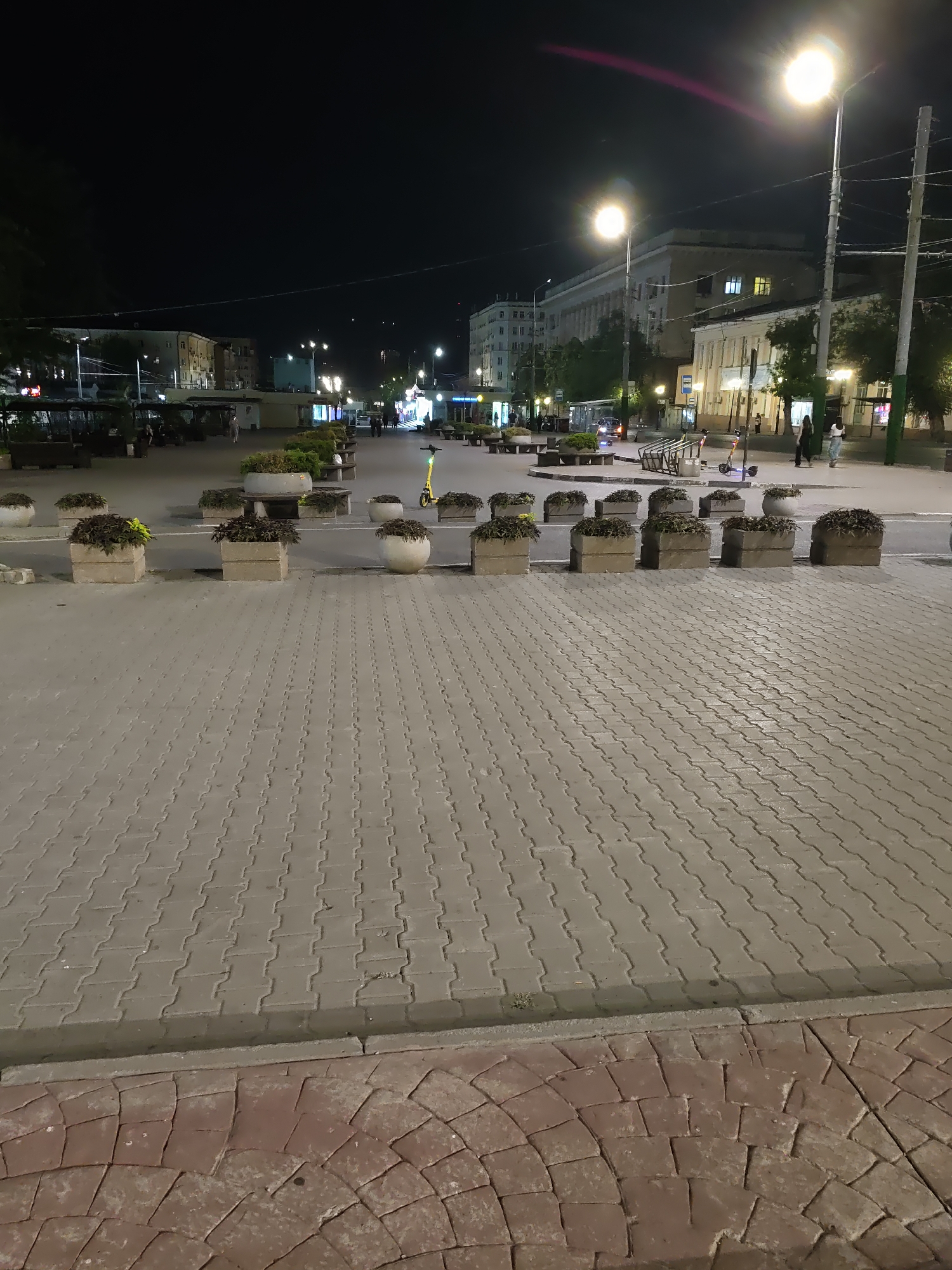 The image depicts a night scene in a city square, likely in Rostov-on-Don, Russia, given the provided context. The foreground is dominated by a paved area with interlocking grey rectangular stones, transitioning to a section with curved, reddish-brown paving stones. In the mid-ground, a street separates the foreground from a line of concrete planters containing dark green foliage. Beyond the planters, benches and more planters are visible, interspersed with streetlights that cast a yellowish glow. Several buildings with illuminated windows line the background, suggesting a commercial or residential area. A few people are seen walking, and a yellow electric scooter is parked near a bike rack. The overall atmosphere is that of a public space at night, with artificial lighting illuminating the scene.