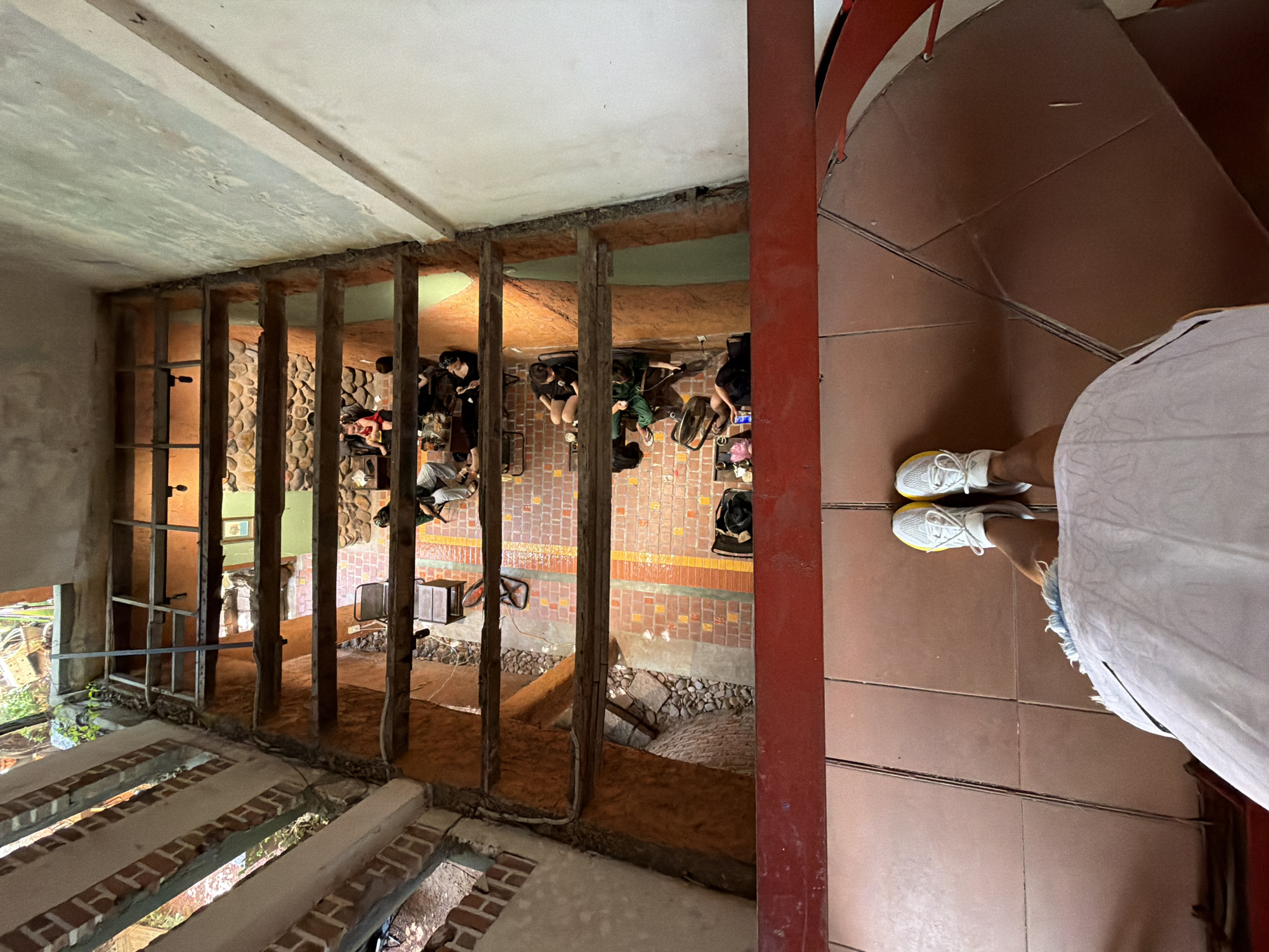 The image shows a view looking down into a multi-level cafe or restaurant in Nha Trang Ward, Vietnam. The architecture features exposed brickwork, wooden beams, and stone accents. Several patrons are seated at tables on different levels, some appearing to be engaged in conversation or working on laptops. The foreground shows the legs of the photographer, wearing white sneakers and a white skirt, standing on a tiled floor near a red railing. The overall impression is that of a casual, somewhat rustic establishment.