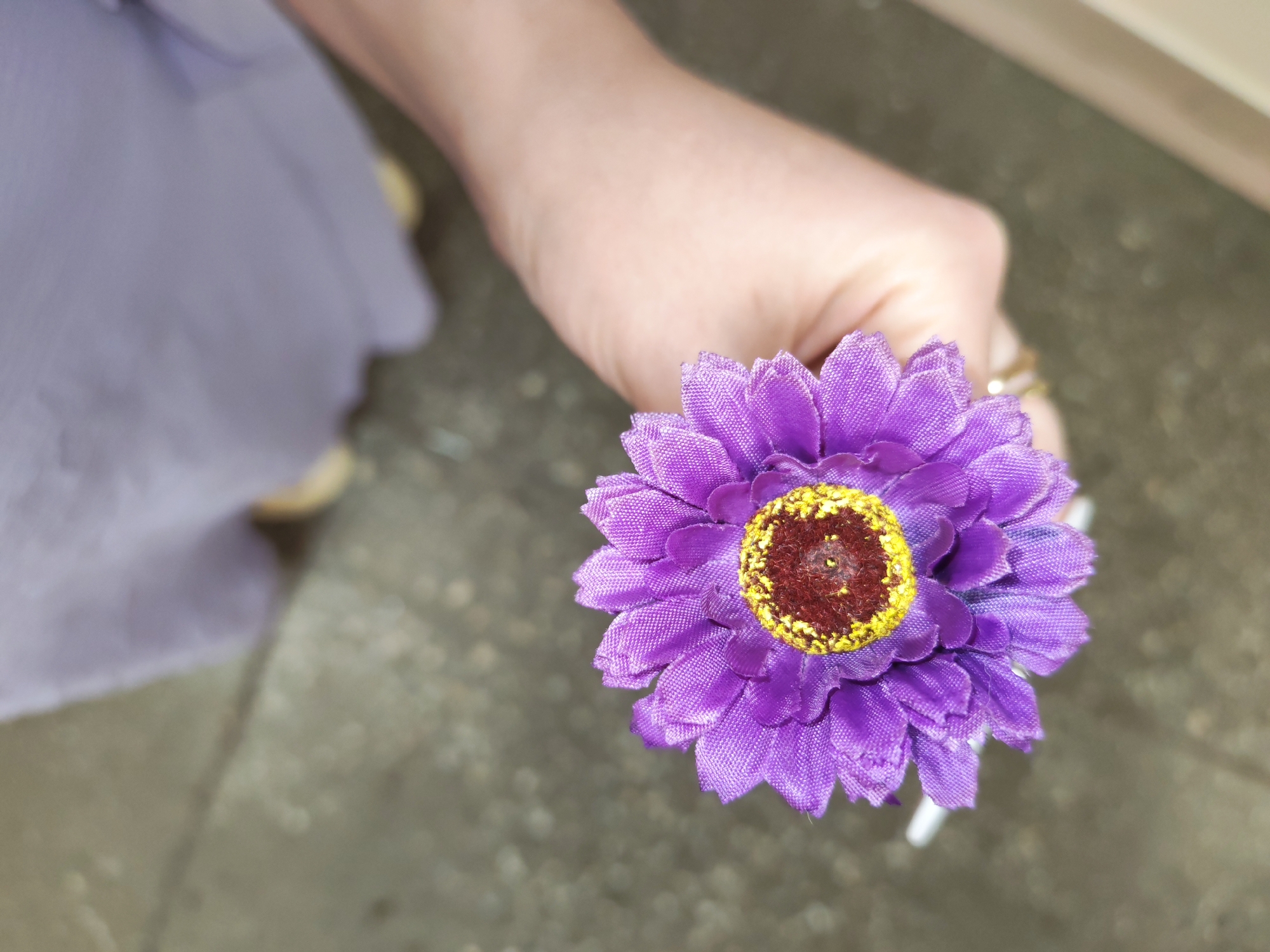 A close-up shot shows a person's hand holding a purple artificial daisy with a yellow and brown center. The person is wearing a light purple garment, possibly a dress or skirt. The background is blurred, showing a gray, textured surface that could be a floor or pavement. The scene appears to be outdoors or in a non-descript indoor location. There is no information in the image to indicate the location is Izhevsk, Russia.