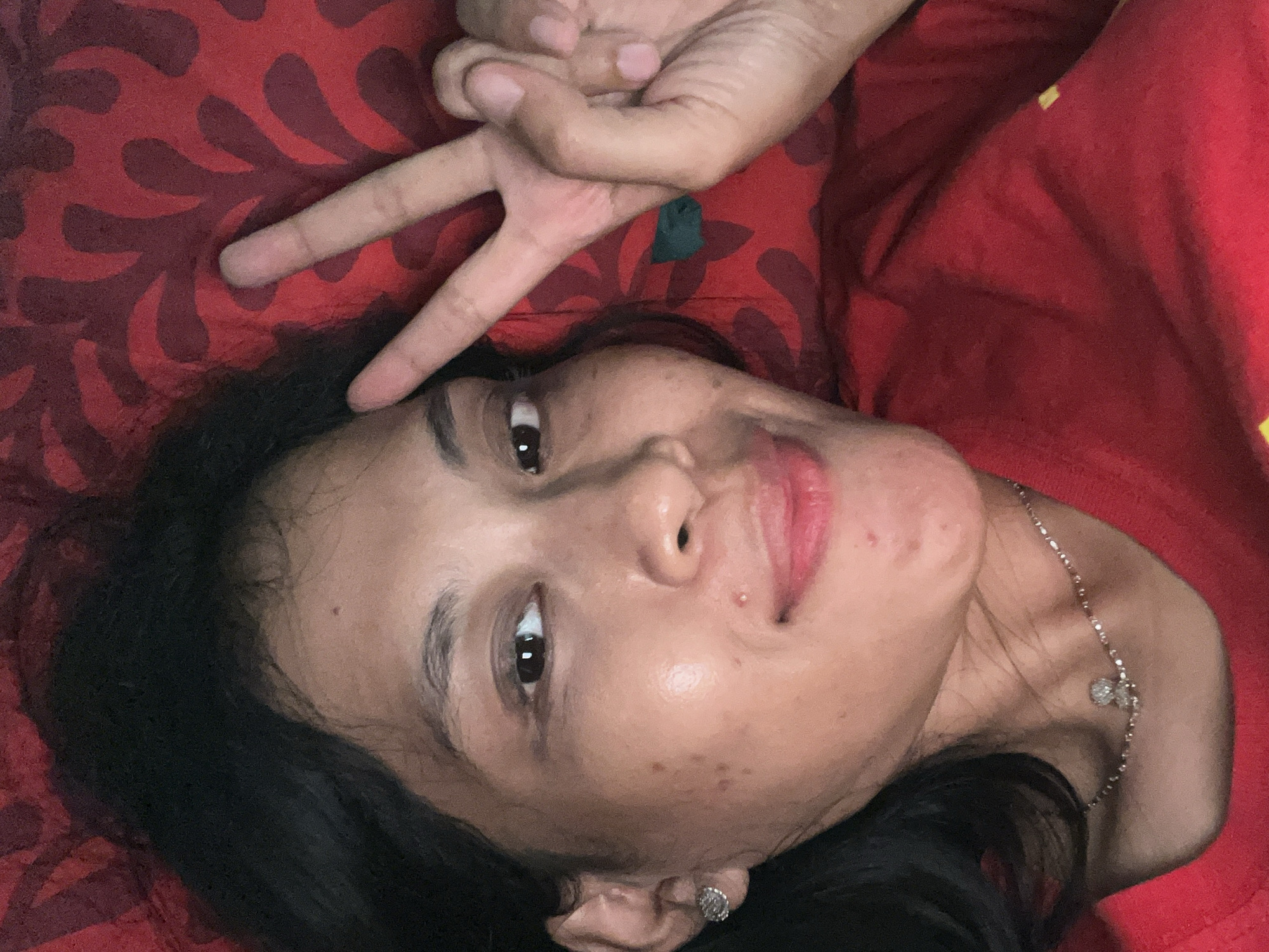 A woman lies on a red surface with a patterned design. Her head is tilted to the right, and she is looking directly at the camera. She makes a V sign with her fingers held above her forehead. She has black hair and is wearing a silver necklace. An earring is visible in her left ear. Her skin has visible blemishes. This scene takes place in Kecamatan Pati, Indonesia.